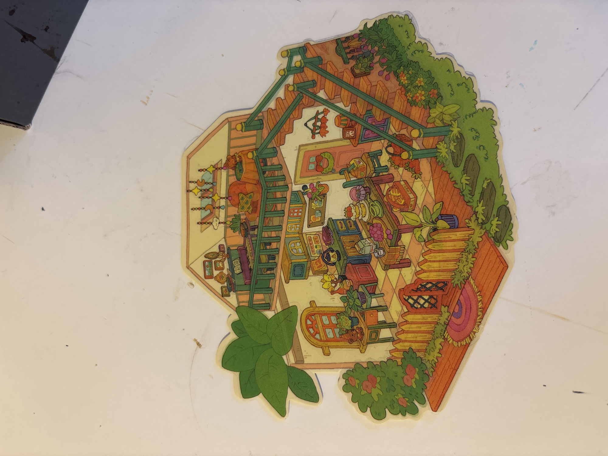 The image is a colorful, cartoon-style illustration of a miniature, multi-story house with a garden. The house appears to be made of wood, with a light yellow exterior and an orange-tiled roof. The interior is visible through the open sides, revealing different rooms and balconies.
The ground floor features a kitchen area with a stove, cabinets, and a table set with food and dishes. There is also a doorway with a wreath and a small table with a lantern. Outside, a wooden fence encloses a garden with a welcome mat, bushes with red flowers, and stepping stones.
The second floor, accessible by an outdoor staircase, includes a living area with a sofa, a table, and a piano. A balcony overlooks the garden. Above this is an attic or upper-level room with a crib-like structure adorned with hanging stars and moons.
The illustration is detailed and whimsical, with small decorative elements like picture frames, plants, and food items. The colors are bright and cheerful, creating a cozy and inviting atmosphere. The overall scene depicts a charming, idealized home.