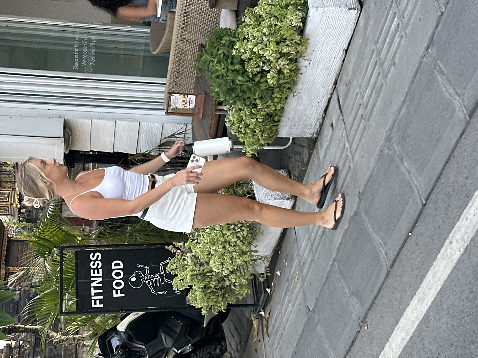 A full-body, medium shot captures a Caucasian woman walking on a tiled sidewalk in front of a building with large glass doors. The woman, with blonde hair pulled back in a bun, is wearing a white tank top, white shorts, and black flip-flops. She carries a white water bottle in her right hand and her left hand is at her side. A black sign with white lettering that reads "FITNESS FOOD" and features a running skeleton illustration is visible to her left, next to a black motorcycle. Green bushes in white planters are positioned behind the sign and to the right of the woman. A sign on a table to the right of the bushes displays images of drinks and the text "30K". The building's glass doors have text printed on them, including "Sleep is for the weak. We're open from 7 am until 11 pm". The environment appears to be a street in North Kuta, Indonesia.