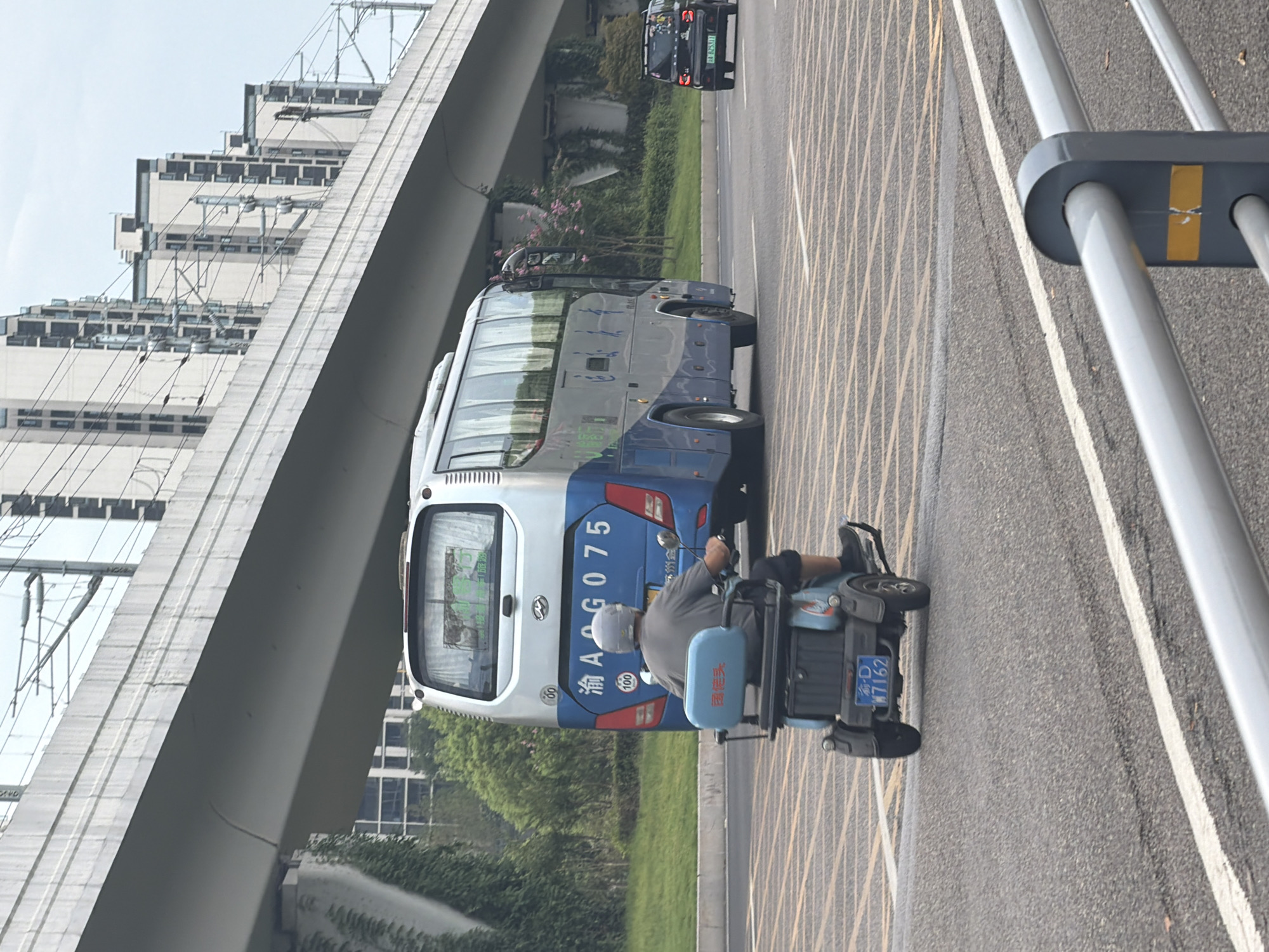 In Jiangjin, China, on a cloudy day, a scene unfolds on a multi-lane road beneath an elevated overpass. The foreground features a grey metal railing with yellow and grey accents. The road surface is asphalt, marked with diagonal yellow lines and a white lane divider.
Dominating the mid-ground is a blue and silver bus with the license plate "渝A AG075". The bus is stationary or moving slowly, facing away from the viewer. To the left of the bus, a person is riding a blue motorized scooter. The rider, wearing a grey shirt and a light grey helmet, is also facing away. The scooter has a blue seat with Chinese characters on the backrest and a license plate that reads "渝D W7162".
In the background, a black SUV with a license plate "渝A 825311" is visible in the right lane, moving away from the viewer. Behind the road, a grassy slope leads up to the elevated overpass, which is a large concrete structure supporting what appears to be a railway or an elevated road. Beyond the overpass, several tall, modern buildings with numerous windows are visible against the overcast sky. Green trees and some flowering bushes are present on the grassy embankment.