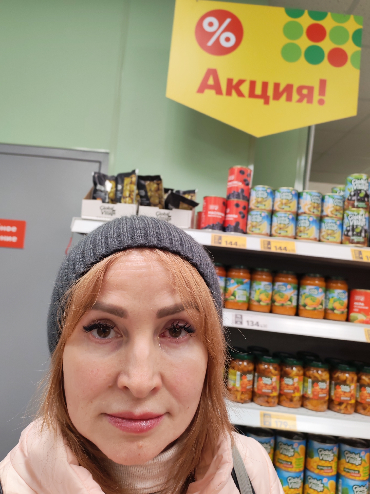 The image captures a close-up selfie of a woman inside a supermarket or grocery store in Chaikovsky, Russia. The woman, appearing to be in her 30s or 40s, has light reddish-brown hair, dark brown eyes, and is looking directly at the camera with a neutral expression. She wears a dark grey knitted beanie, a light pink puffer jacket, and a light-colored turtleneck sweater. Her left eye (viewer's right) shows some redness or irritation.
Behind the woman, a retail environment is visible, characterized by multiple metal shelves stocked with various food products. Products include black packaged items on an upper shelf, numerous cans with blue, yellow, and red labels (some branded "Global Village"), and several glass jars containing orange-colored pureed or sauced items. Price tags are visible on the shelves, with one reading "134" and another "179" in a yellow rectangular tag.
Above the shelves, a prominent yellow, hexagonal sign hangs, displaying a red circle with a white percentage symbol and the red Cyrillic word "Акция!", which translates to "Promotion!" or "Sale!". To the far left, a light green wall and part of a grey door or wall are visible, with a partially obscured red rectangular sign that appears to contain Cyrillic text. The overall lighting is consistent with indoor artificial retail illumination.