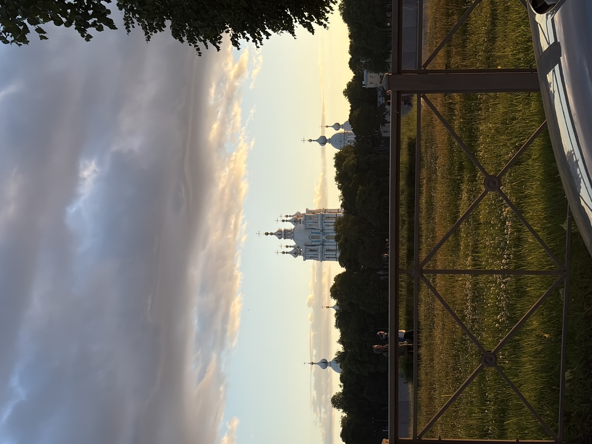 The image depicts the Smolny Cathedral in Saint Petersburg, Russia, at dusk. The sky is filled with dramatic, cloudy formations that are illuminated by the setting sun, creating a warm, golden glow on the horizon. In the foreground, a metal fence with a criss-cross pattern encloses a grassy field dotted with white wildflowers. Two individuals are visible walking in the distance, silhouetted against the bright sky. Beyond the field, a line of dark green trees serves as a backdrop for the iconic white and blue spires of the Smolny Cathedral complex, which stands prominently against the illuminated sky. Part of a car's hood is visible in the bottom right corner of the frame, suggesting the photograph was taken from a street or walkway.