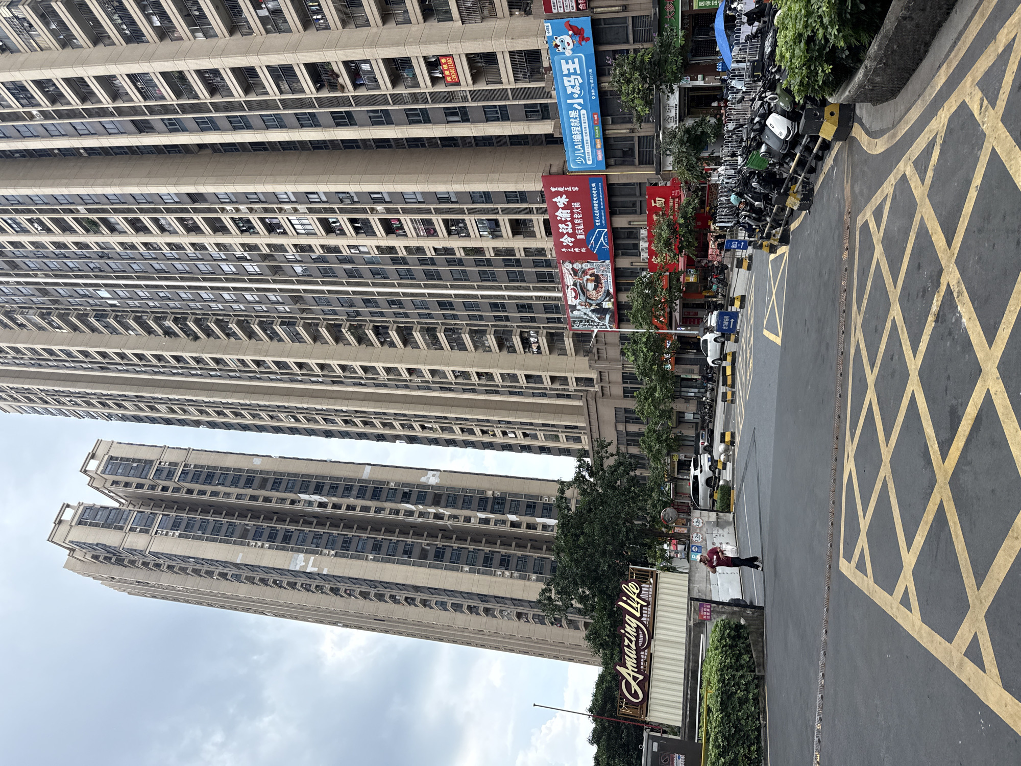 The image captures a street scene in Haizhu District, China, dominated by two towering, modern apartment buildings. The sky is overcast. In the foreground, a wide asphalt road features prominent yellow crisscross markings indicating a no-parking zone. A few vehicles, including motorcycles and cars, are parked along the right side of the road. A person in a red shirt and dark pants is walking away from the camera on the sidewalk near the entrance to a building complex. Signs with Chinese characters are visible on the buildings, along with advertisements. Greenery, including trees and bushes, adds a touch of nature to the urban environment. The architecture suggests a dense residential area with numerous balconies and windows on the apartment blocks.