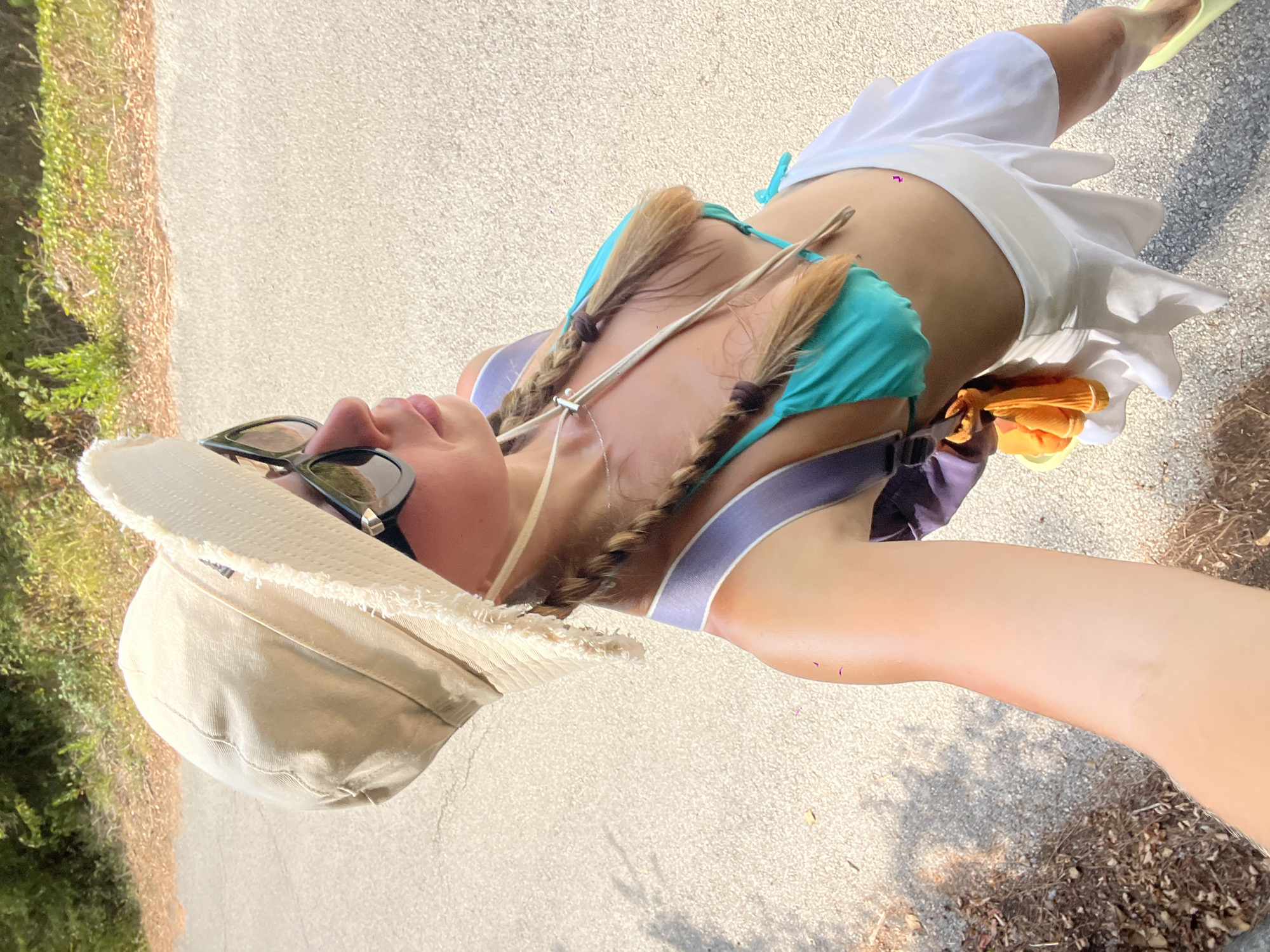 A person stands on a paved road in an outdoor environment. They are wearing a light-colored wide-brimmed hat, sunglasses, a turquoise bikini top, and a white skirt. The person has braids and is carrying a backpack. The background consists of dry vegetation and a paved surface. The lighting suggests it is daytime.