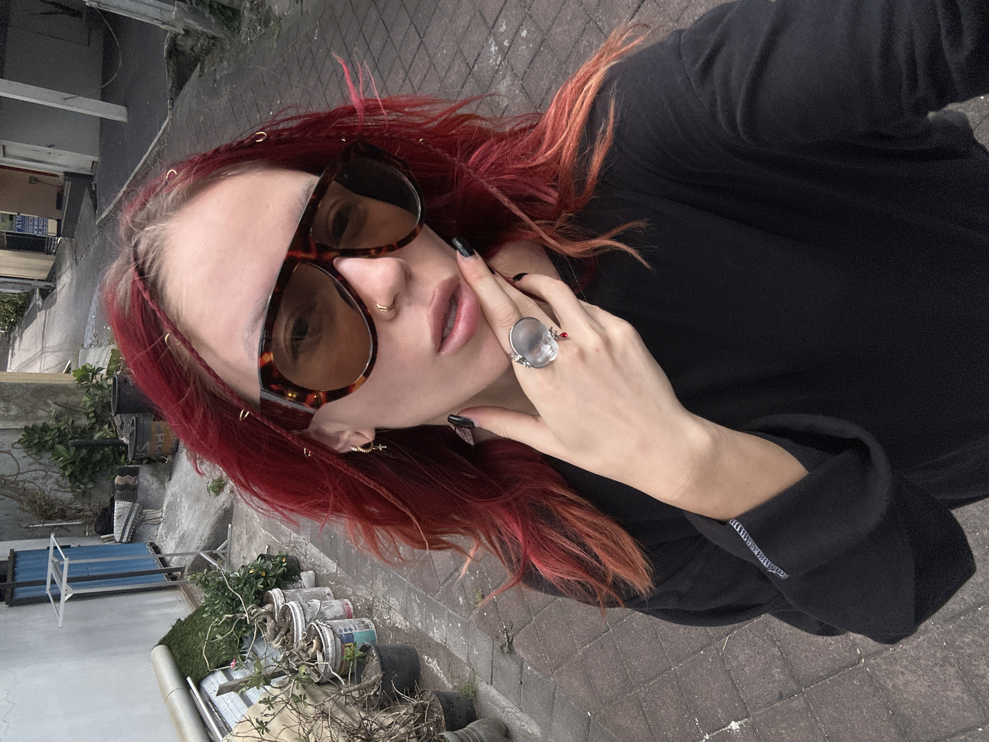 A medium shot captures a person with vibrant red hair, styled in braids and loose strands, taking a selfie on a street in North Kuta, Indonesia. The individual wears large, tortoise-shell sunglasses, a nose ring, and a black long-sleeved shirt. Their left hand, adorned with a skull-shaped ring with a red accent and black nail polish, is raised to their face, touching their chin. The background shows a narrow, paved alleyway with buildings and potted plants lining the sides. The ground is paved with interlocking bricks, and some debris is visible. The overall lighting suggests an overcast day.