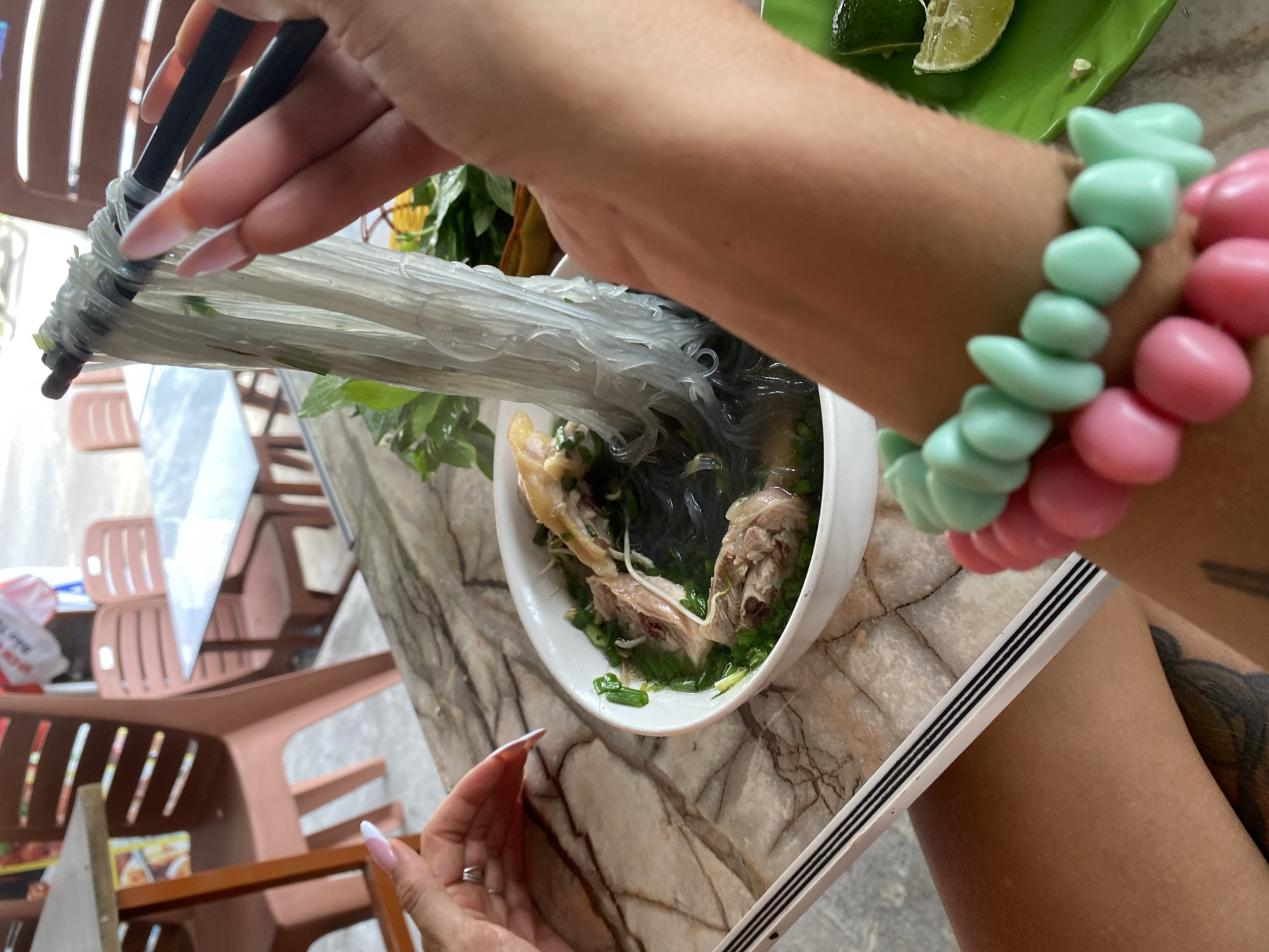 A first-person perspective captures a person actively eating a bowl of Vietnamese noodle soup, likely Miến Gà (chicken glass noodle soup), in what appears to be an informal eatery in Tien Thanh Ward, Vietnam.
The dominant feature is a hand, seen from the wrist down, holding a pair of black chopsticks and lifting a large, entangled portion of translucent glass noodles from a white bowl. The hand sports long, light pink-tipped acrylic nails. On the wrist, two chunky, colorful bracelets are visible: one mint green and the other pink, adding a playful touch. Another hand, adorned with a silver ring on the index finger, is partially visible at the bottom left, resting near the bowl. A prominent, dark-colored tattoo is partially visible on the upper thigh or leg, suggesting the person is wearing shorts or a skirt.
The noodle soup contains bone-in chicken pieces, possibly wings or thighs, along with ample amounts of fresh green herbs like chopped scallions and potentially mint or basil, all submerged in a clear broth. To the left of the bowl, a small green plate holds several lime wedges, a common condiment for Vietnamese soups.
The meal is served on a table with a distinct black and white marble-like pattern. In the background, several brown plastic chairs, some stacked, suggest a casual, possibly outdoor or semi-outdoor dining environment. Natural daylight illuminates the scene, indicating it is daytime. A blurry red and white plastic bag with indistinct text can be seen in the distant background.