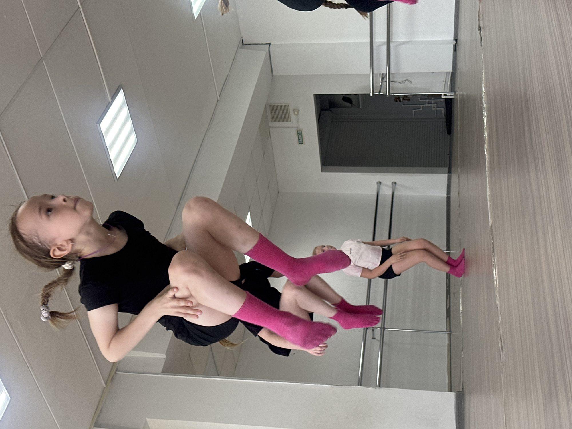 Two young girls are in a dance studio in Votkinsk, Russia. The girl in the foreground is wearing a black t-shirt, black shorts, and pink socks; her blonde hair is in pigtails. She is sitting and looking upwards. Behind her, another girl is wearing a white shirt, black shorts, and pink socks. She is lying down, also looking upwards. The studio has a gray wall with a mirror that reflects both girls, creating a sense of multiple individuals. Behind them, there are metal ballet barres. The ceiling features rectangular light panels. A gray door is visible in the background.