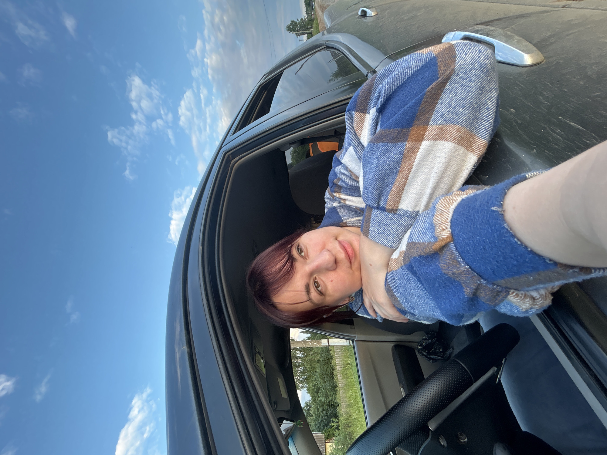 In the city of Fokino, Russia, a young woman with short, dark hair is seen from the chest up, leaning out of the open window of a car. She is wearing a blue, white, and brown plaid shirt. Her arms are crossed, and her head is resting on her arms. Her gaze is directed upwards and to the right, with a slight smile. The car's interior, including a steering wheel, is visible on the left side of the image. Outside the car, trees and a wooden fence can be seen, suggesting a rural or semi-rural setting. The sky above is blue with scattered white clouds.