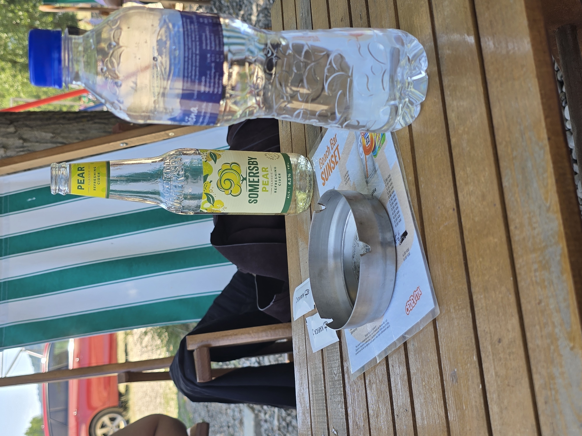 A medium shot shows a wooden table at an outdoor venue in Zvornik, Bosnia and Herzegovina. On the table are a bottle of Somesrby Pear refreshing cider, a plastic bottle of water with a blue cap, an ashtray, and a menu. In the background, a red car is parked near a green and white striped deck chair. The ground is covered with gravel.