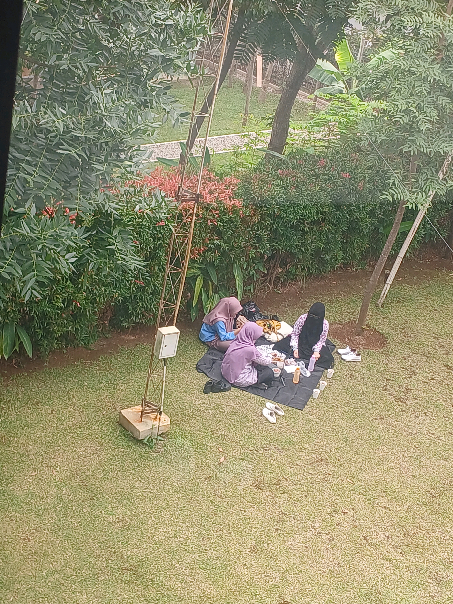 The image depicts a picnic scene in a grassy area, likely within the city of Arjasari, Indonesia, surrounded by lush green trees and vegetation. In the center of the frame, three individuals, all wearing hijabs, are seated on a black picnic blanket. They are engaged in a meal, with food and drinks spread out before them. To the left, one person in a light purple hijab is reaching for food, while another in a blue hijab is looking down at the spread. To the right, a third individual wearing a black niqab is seated, holding a bottle and a cup. Several white cups and a brown bottle are visible on the blanket. Two pairs of white sandals are placed on the grass, one near the picnic blanket and another further to the right. A metal tower, possibly an antenna or utility pole, stands to the left of the picnic, with a white box attached to its structure. The ground is covered in short, well-maintained green grass. The background is filled with dense green foliage and trees, suggesting an outdoor setting, possibly a park or a backyard.