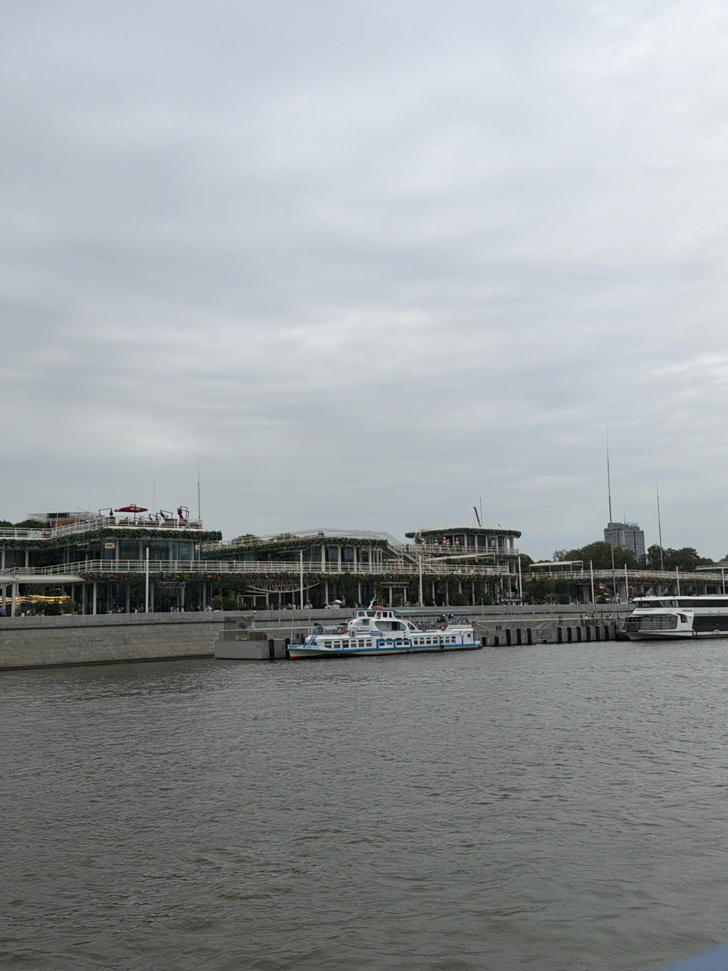 This image captures a view of the Patriarchs' Pontoon on the Moskva River in Moscow, Russia. A multi-story building with a modern architectural design, featuring extensive glass windows and white railings, lines the riverbank. The building is adorned with greenery and decorative elements. Several boats are docked along the pontoon. In the foreground, a passenger boat with blue and white detailing is moored. To its right, another larger passenger boat, predominantly white with dark windows, is also docked. The river is a murky gray color with visible ripples on its surface. The sky is overcast with a thick layer of gray clouds. In the background, trees and a tall building are visible.