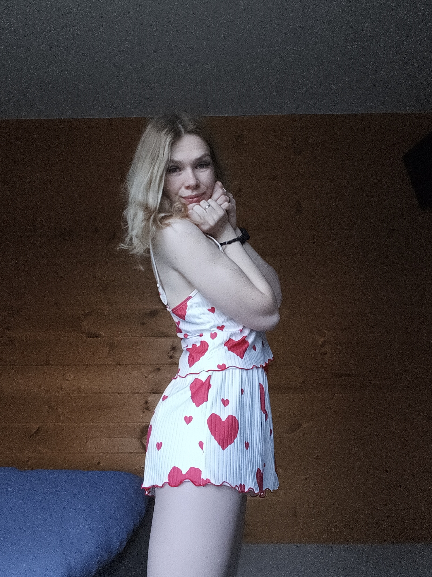 A young, fair-skinned woman with shoulder-length blonde hair is posing indoors. She is wearing a white dress with red heart patterns. Her hands are clasped near her face. A blue cushion is visible on a bed near the woman, and the background consists of a wooden wall and a dark ceiling. The image does not contain sufficient information to pinpoint the precise location within Русский Вожой, Russia.
