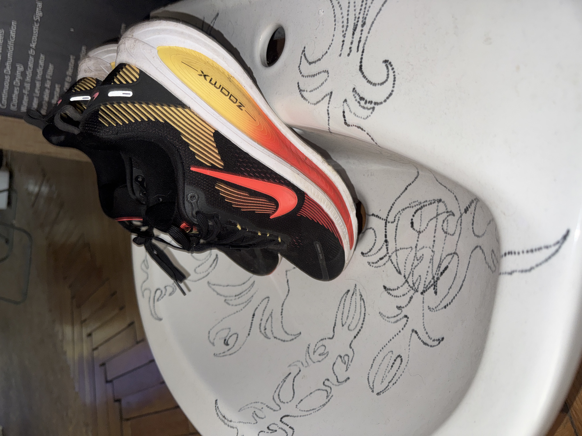 A pair of black, red, and gold athletic shoes, identified as Nike ZoomX Invincible Run Flyknit 3, is resting inside a white ceramic basin. The shoes feature a prominent red Nike swoosh on the side and "ZOOM X" written in yellow on the white midsole. The basin has distinctive abstract, wavy patterns composed of dotted black lines drawn or painted on its interior surface. To the left of the basin, a dark appliance panel with white text is partially visible, displaying phrases such as "Continuous Dehumidification," "(Clothes Drying)," "Water Level Indicator," "Water-Full Indicator & Acoustic Signal," "Washable Air Filter," and "Restart." Below the basin, a light brown wooden floor is visible. The scene is indoors, likely a utility room or bathroom, with artificial lighting. There are no people or active events in the image. The location context is Sofia, Bulgaria.