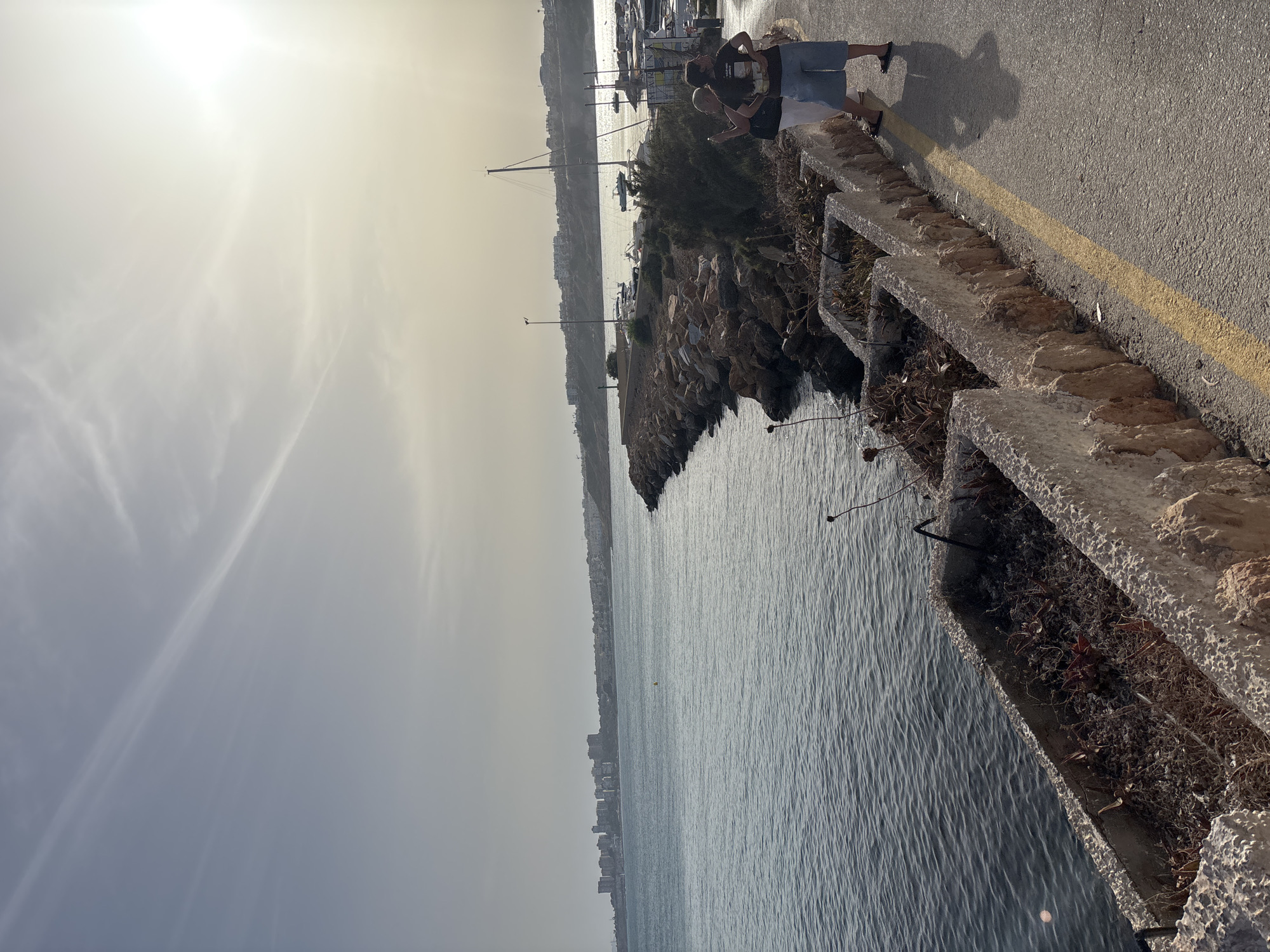 The image depicts a coastal scene in Urbanización Cabo Roig, Spain. In the foreground, a paved pathway with a yellow line runs alongside a concrete planter box filled with dry vegetation. Two individuals, one wearing a dark t-shirt and denim shorts, and another in a white top and light-colored pants, stand on the pathway facing away from the camera, seemingly interacting. Behind them, a rocky breakwater extends into the sea, lined with various boats docked in a marina. The sea itself is calm, reflecting the overcast sky. In the distance, a city skyline with numerous buildings stretches along the coast under a hazy, sunlit sky. The overall environment suggests a warm, possibly late afternoon or early morning setting.