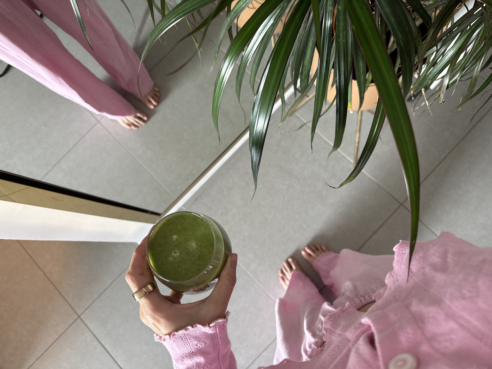 A top-down shot depicts a person in pink attire holding a green smoothie in a glass. The person's feet are bare. A plant with long, green leaves is positioned on the right side of the frame. A mirror reflects a portion of the person's body and legs, creating a visual doubling effect. The floor is tiled in a light gray color with a grid pattern. The location is in Ramat Gan, Israel.