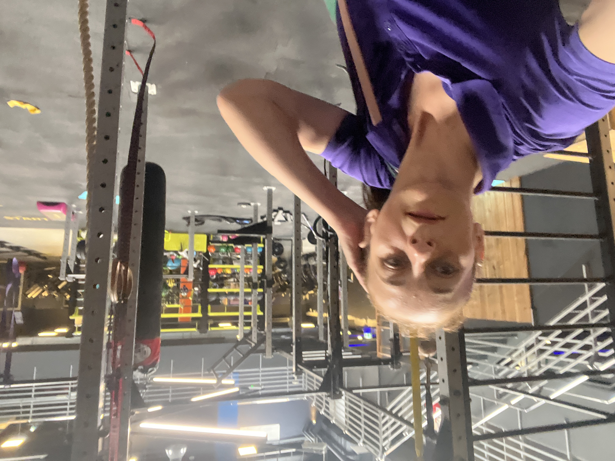 An indoor, eye-level, medium shot captures a woman with blonde hair, wearing a purple polo shirt, in a gym. She is facing the camera and touching her hair with her right hand. Her left arm is bent at the elbow. The gym is equipped with various exercise machines, including a punching bag, weight racks, and a multi-level structure with bars. The floor is dark grey. The lighting is bright, with fluorescent lights visible on the ceiling. The background includes wooden accents and metal gym equipment.
