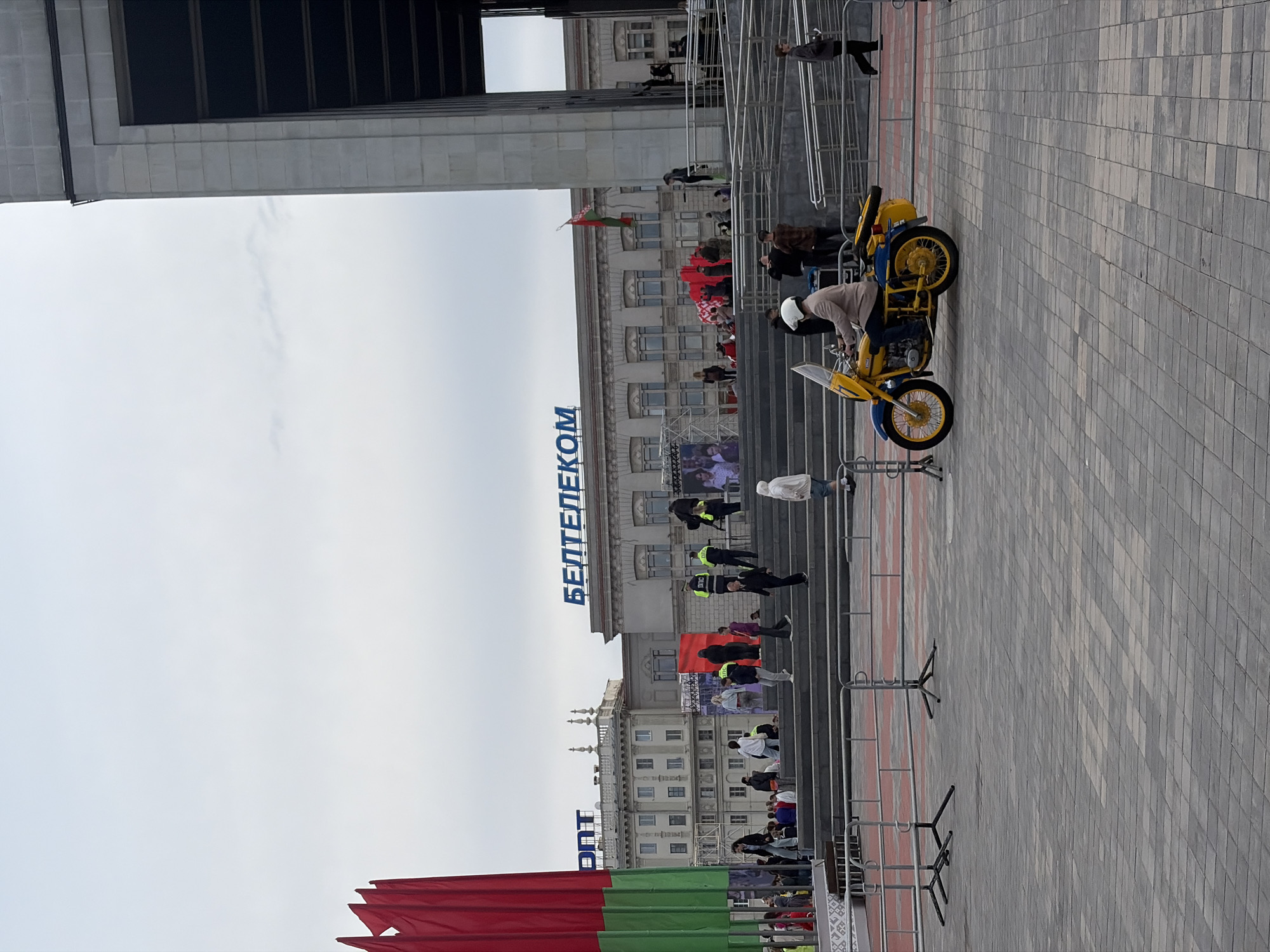 The image depicts an outdoor scene in Minsk, Belarus, near the central entrance of the Beltelecom building. The foreground is dominated by a gray tiled pavement and metal barricades. A yellow and blue motorcycle with a rider wearing a beige jacket and white helmet is positioned on the pavement, facing right. In the mid-ground, a set of wide, grey concrete stairs leads up to the Beltelecom building. Various individuals are seen walking on these stairs and gathered at the top. Behind the stairs, the Beltelecom building is visible, featuring large blue lettering spelling out "БЕЛТЕЛЕКОМ". To the left of the Beltelecom building is another ornate building with light-colored facades and decorative elements. A large red and green flag, the national flag of Belarus, is prominently displayed on the left side of the image, partially obscuring the view of the buildings. The sky is overcast, suggesting a cloudy day. Security personnel in yellow vests are visible among the crowd on the stairs. The overall scene suggests a public event or gathering.