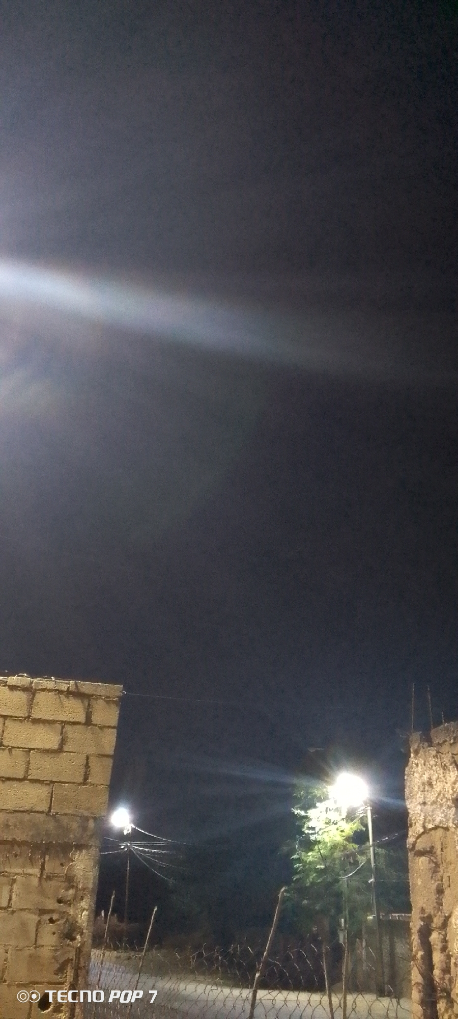 The image depicts a nighttime scene in Altagracia, Venezuela. In the foreground, there is a chain-link fence. Behind the fence, there are two walls constructed from stone and cinder blocks. Streetlights illuminate the area, and power lines run between the light poles. Vegetation is visible in the background, likely trees or shrubs. The sky is dark and streaked with light. The "TECNO POP 7" watermark is visible in the lower-left corner of the image.