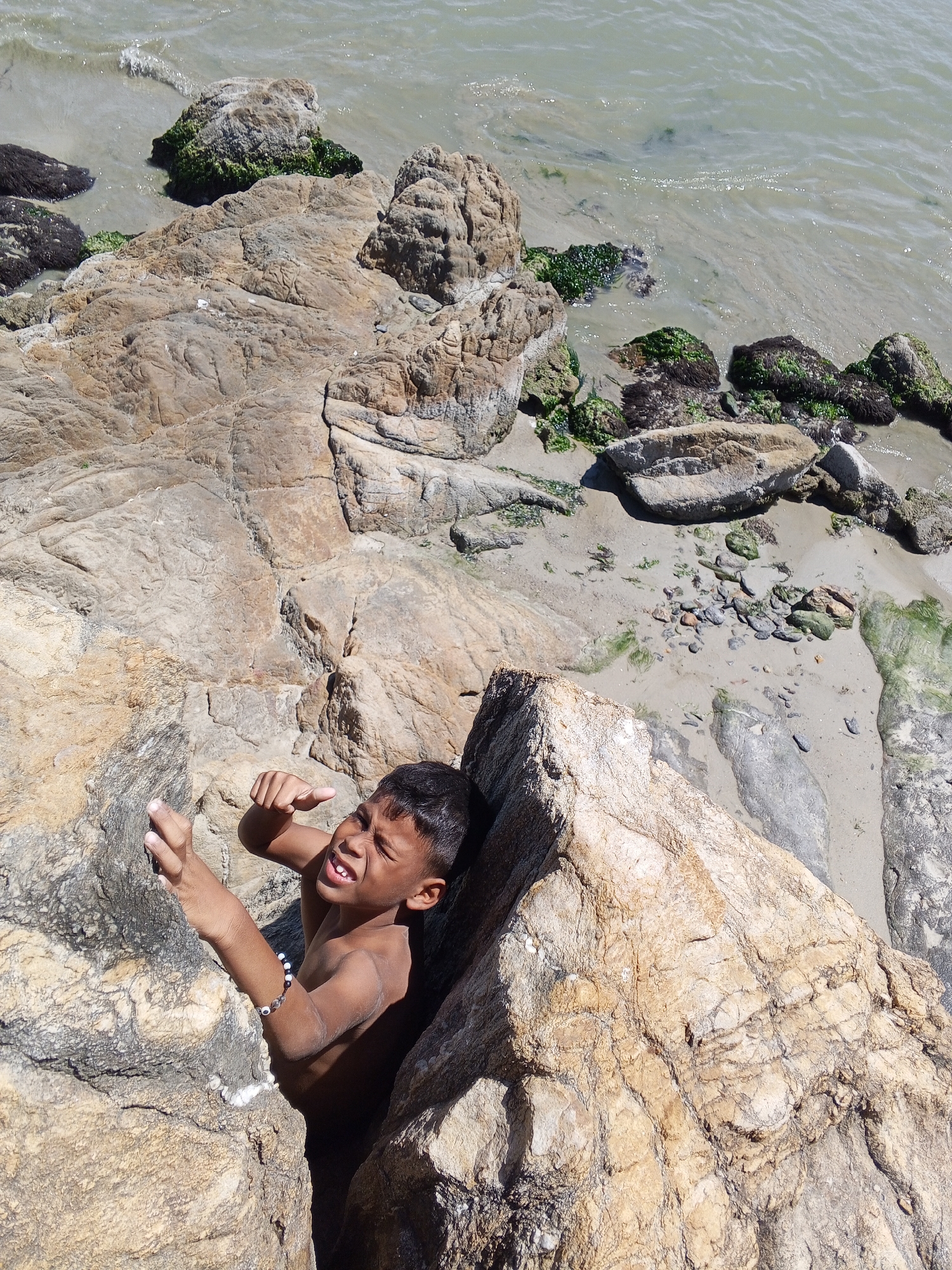 A young boy is wedged between two large rock formations at a beach in Juan Griego, Venezuela. The rocks are a light brown color with intricate patterns and crevices. To the boy's left, there are several other rocks, some covered in green algae, leading towards the ocean. The water is a murky gray-green color. The boy is shirtless and wearing a beaded bracelet on his left wrist. He is looking up with a slightly pained expression, his right hand raised in a fist, and his left hand reaching towards the rocks to his left. The sand in between the rocks is a light beige color with small pebbles scattered around. The overall lighting is bright, suggesting a sunny day.