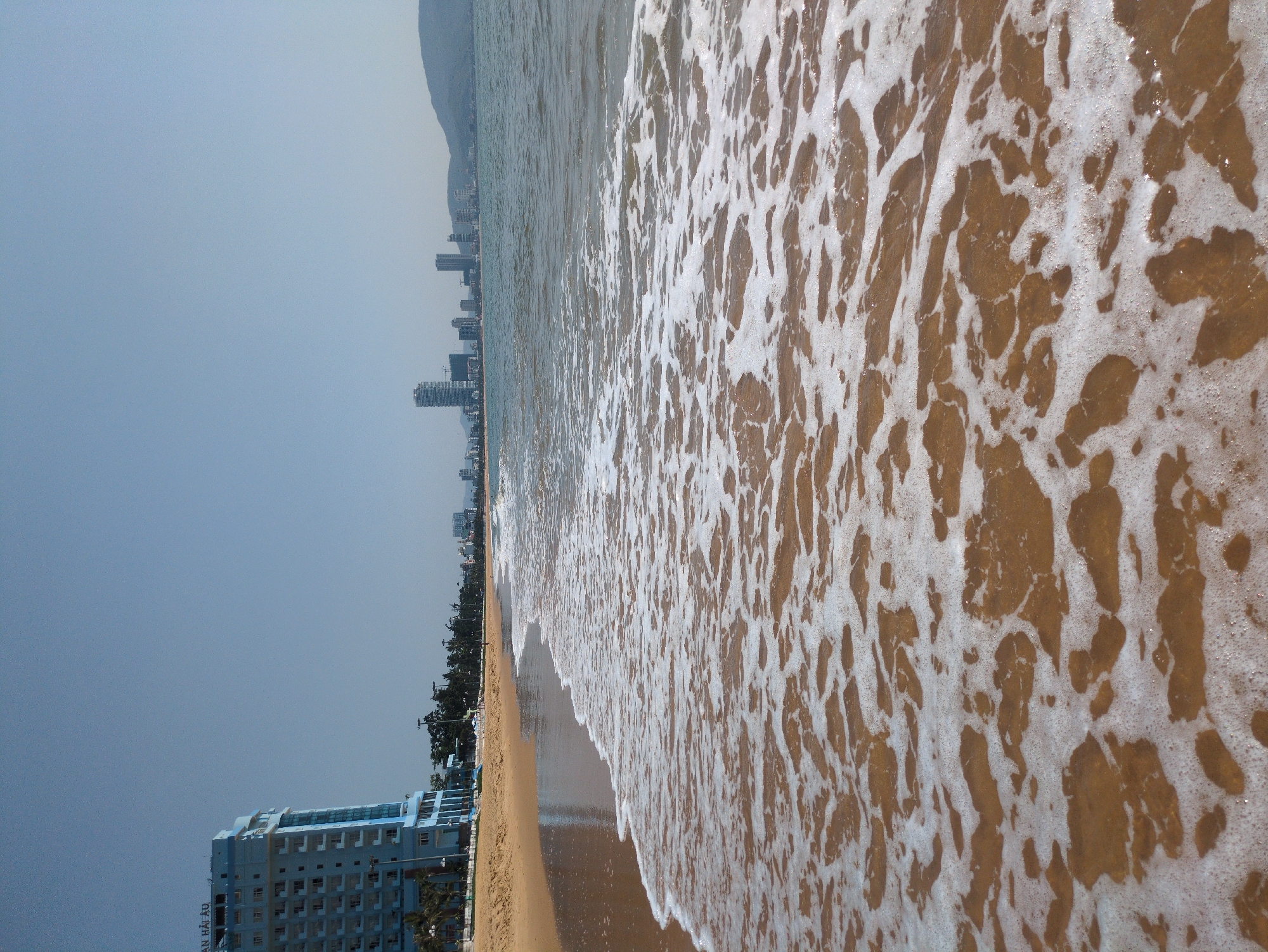 The image shows a beach in South Quy Nhon Ward, Vietnam. Waves are washing up on the sandy shore. In the background, a line of buildings is visible along the coastline, with a mountain in the distance. A taller building is visible in the bottom left corner of the image. The sky is a pale blue.