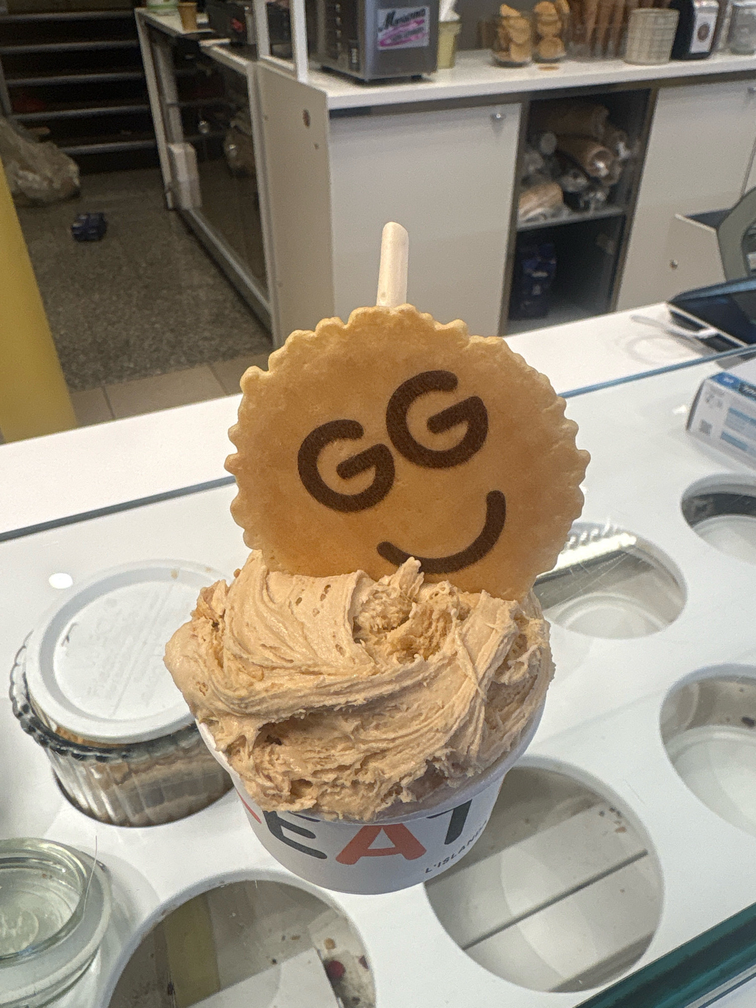 An ice cream cone decorated with a smiling face made of cookies sits on top of a cup of ice cream. The ice cream is a light brown color and has a swirl pattern. The cookie is round and has a ridged edge. The smiling face is made of two dark brown circles for eyes and a curved line for a mouth. The cup the ice cream is in has the word "EAT" printed on it in large letters. The scene appears to be in an ice cream shop. The background shows shelves with various items on them, as well as a counter and a staircase. There are no people visible in the image. The image is taken from a slightly high angle, looking down at the ice cream.