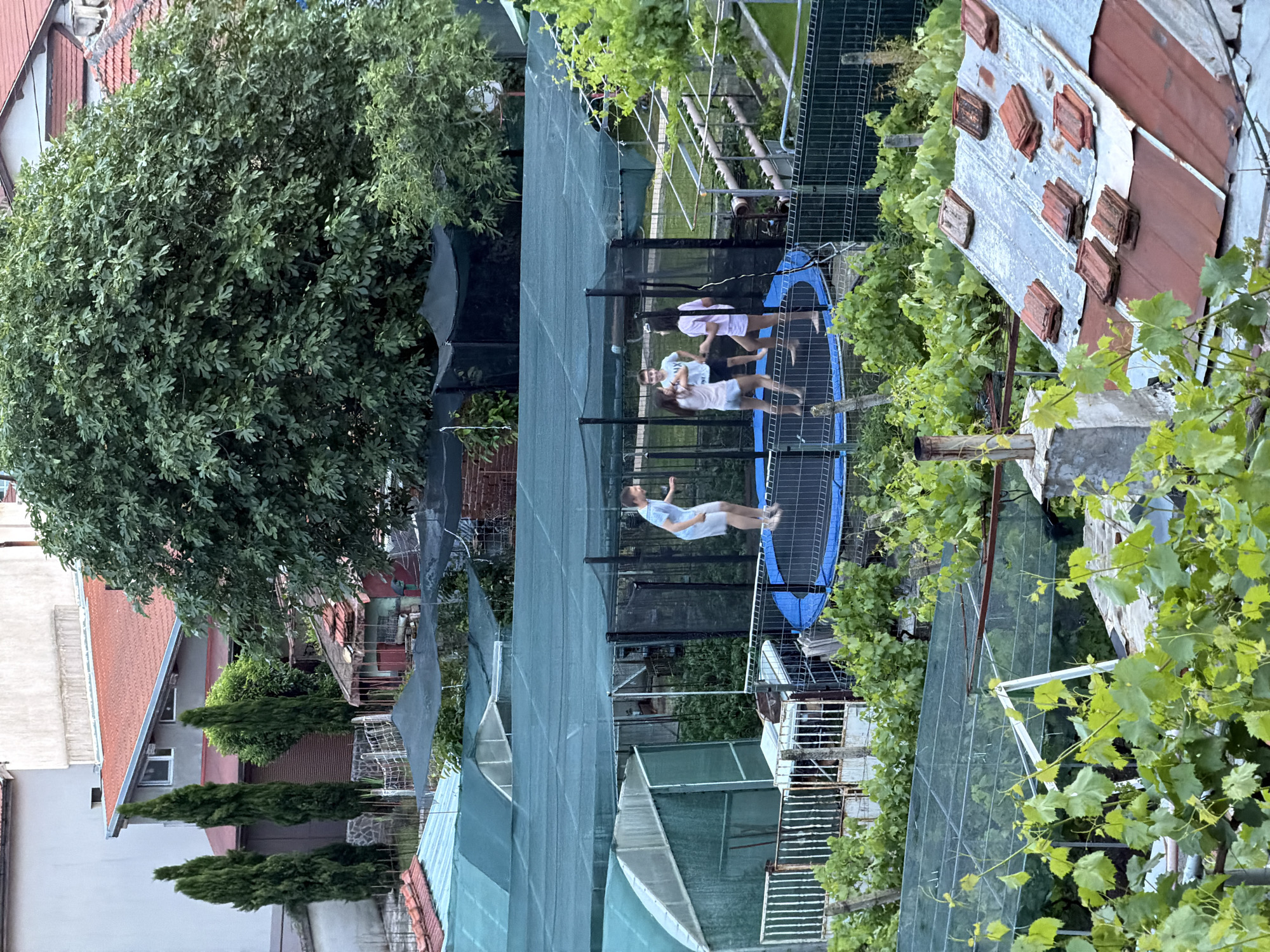 Captured from a high vantage point in Burgas, Bulgaria, the scene focuses on a backyard activity area. Three individuals are visible on a trampoline enclosed by a safety net. One person is airborne, jumping. A green canopy covers an adjacent area, likely a garden or outdoor space. The surroundings include residential buildings with red-tiled roofs and greenery, possibly vines or trees, obscuring some of the structures. A weathered, partially dilapidated building with a chimney is also visible. The photo appears to be taken in the late afternoon or early evening, given the lighting conditions.