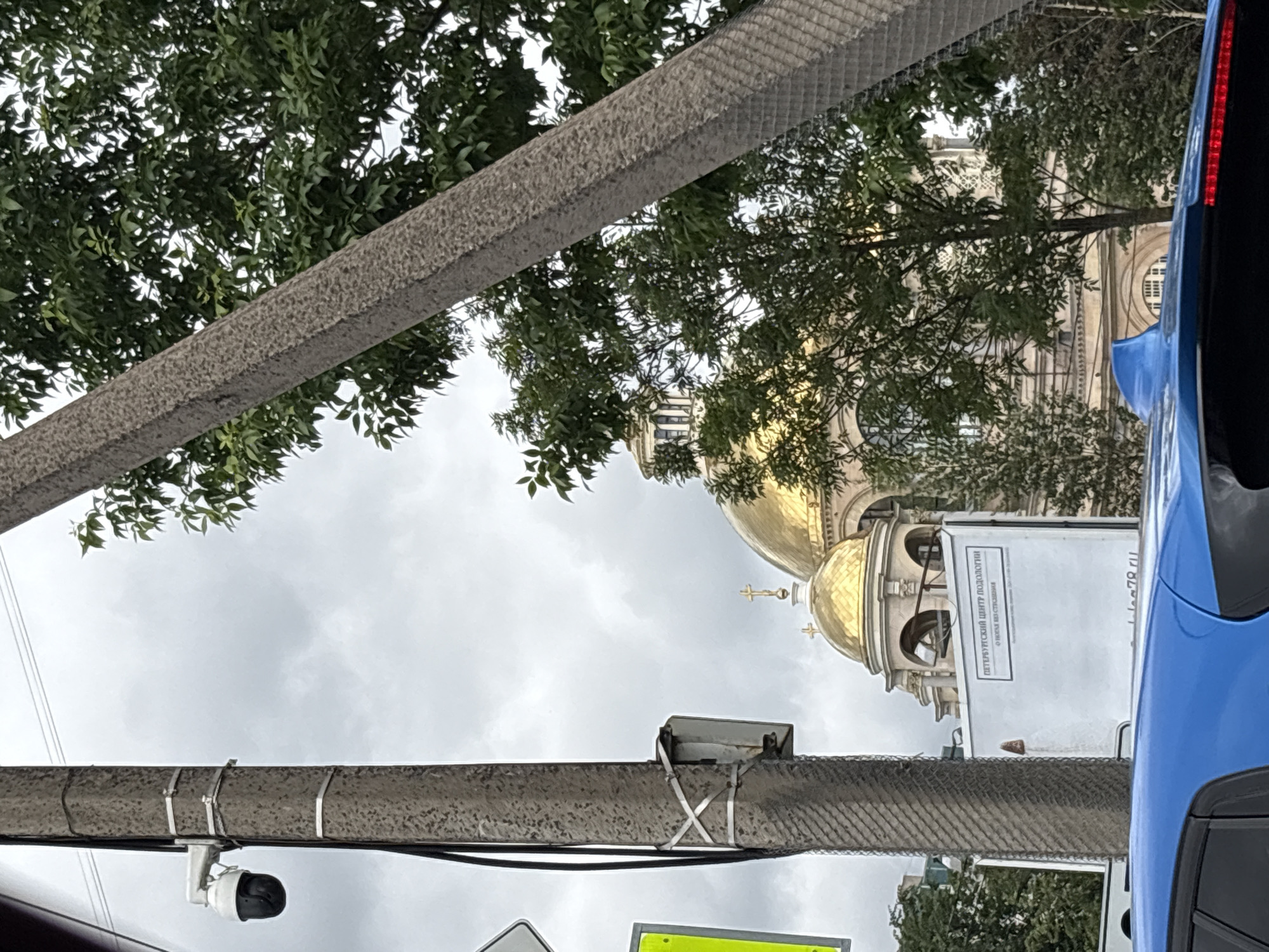 The image is an outdoor, slightly tilted, medium shot featuring a street scene in Saint Petersburg, Russia. The primary subjects are architectural elements of a building with golden domes and a blue car. In the foreground, a concrete structure, possibly a barrier or bridge, runs diagonally across the frame, partially obscuring the view. Attached to this structure is a white security camera. Above, green foliage from trees drapes over the scene.
The background is dominated by a grand building with multiple golden domes, one of which is prominently visible. This architectural style is characteristic of Orthodox churches. A cross is visible atop one of the domes. To the right of the domes, a white rectangular sign with text is attached to the building. The visible text on the sign appears to be in Cyrillic and includes the words "ПЕТЕРБУРГСКИЙ ЦЕНТР ПОДОЛЬИН" and "О НОВЫХ ВЕЩАХ". Below this, a website address "lea.78.ru" is partially visible.
A portion of a blue vehicle is on the far right of the frame, suggesting the photo was taken from inside or alongside a car. The sky is overcast and cloudy, indicating a possibly cool or temperate day, typical of Saint Petersburg. The overall scene suggests an urban environment with a mix of modern infrastructure (car, camera) and historical architecture. There are no people visible in the frame, and no specific activity or event is taking place; it appears to be a still moment captured while passing by. The lighting is diffused due to the cloudy conditions.
