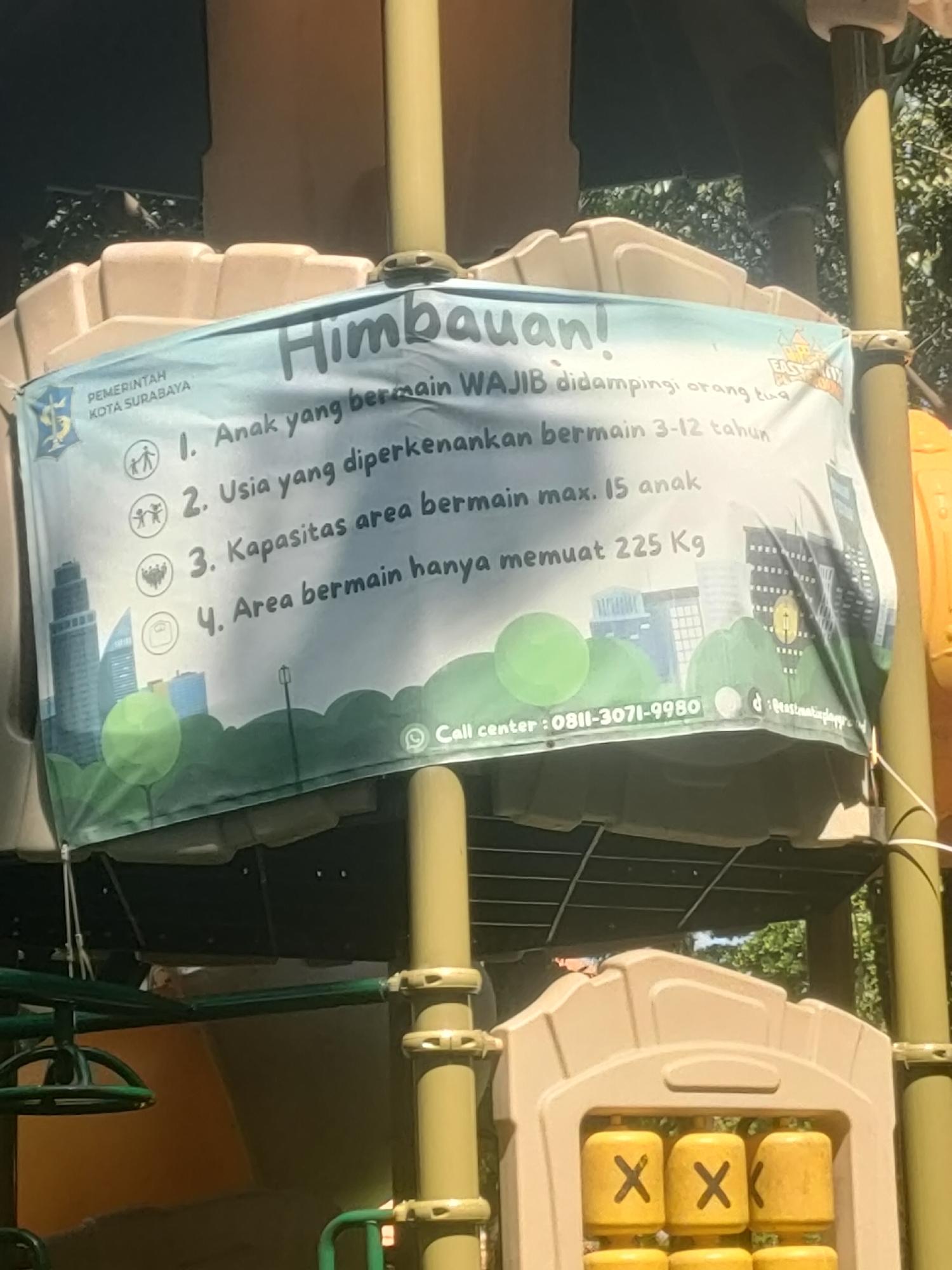 The image shows a banner on a playground structure in Surabaya, Indonesia. The banner, written in Indonesian, displays regulations for the playground, including age limits and capacity restrictions. The top of the banner reads "Himbauan!" (Announcement!). Below this, it lists four points: 1. Children playing must be accompanied by a parent/guardian. 2. Children aged 3-12 years are allowed to play. 3. The area can accommodate a maximum of 15 children. 4. The play area can hold a maximum of 225 kg. There are icons next to each point representing the regulation. The banner features graphics of buildings and trees, and a call center number is provided. In the foreground, the yellow structure of a children's game with X's and O's is visible. The background consists of trees and part of a playground structure.