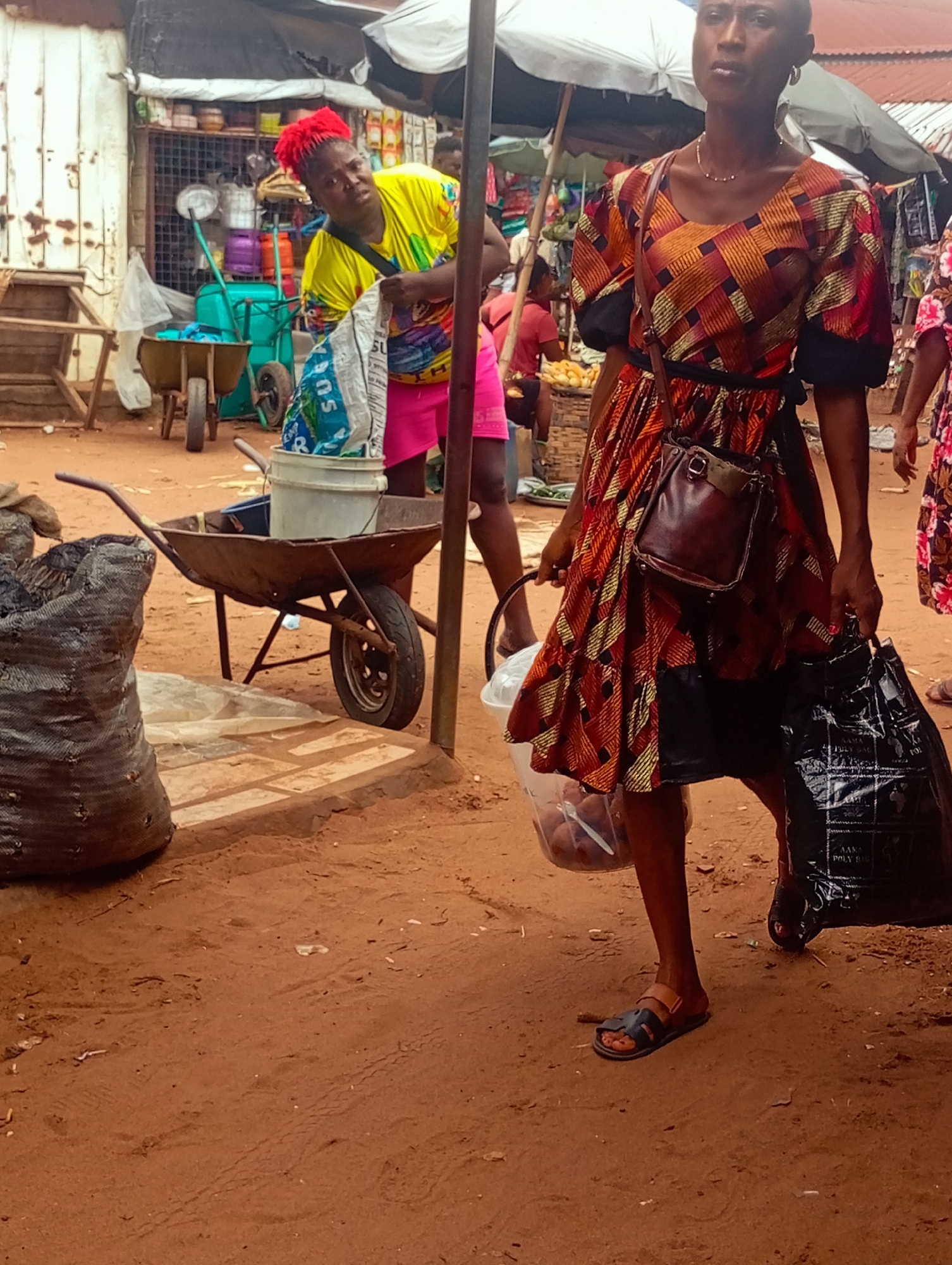 This media file depicts an outdoor market scene. The primary subjects are two women. The woman in the foreground is wearing a vibrantly patterned dress in shades of orange, red, and yellow, with puffed sleeves. She has a small brown leather satchel slung across her body and is carrying a clear plastic bucket filled with what appear to be produce, and a large black plastic bag in her right hand. She is wearing dark sandals.
The second woman, in the background, is wearing a bright yellow t-shirt and pink shorts. She has striking red hair and is looking to the side. She appears to be standing near a cart and some shelves laden with goods, possibly food items.
The setting is an open-air market with stalls, umbrellas providing shade, and sandy ground. There are various objects scattered around, including a wheelbarrow filled with a white bucket and a blue container, large sacks, and more produce displayed in baskets. The overall atmosphere suggests a busy market day. The lighting indicates it is daytime, likely during daylight hours. The presence of produce and market stalls implies a commercial activity where goods are being bought and sold. There is text visible on the black plastic bag the woman in the foreground is carrying, which appears to be a brand or type of bag, possibly "POLY BAG" and "MADE IN NIGERIA".