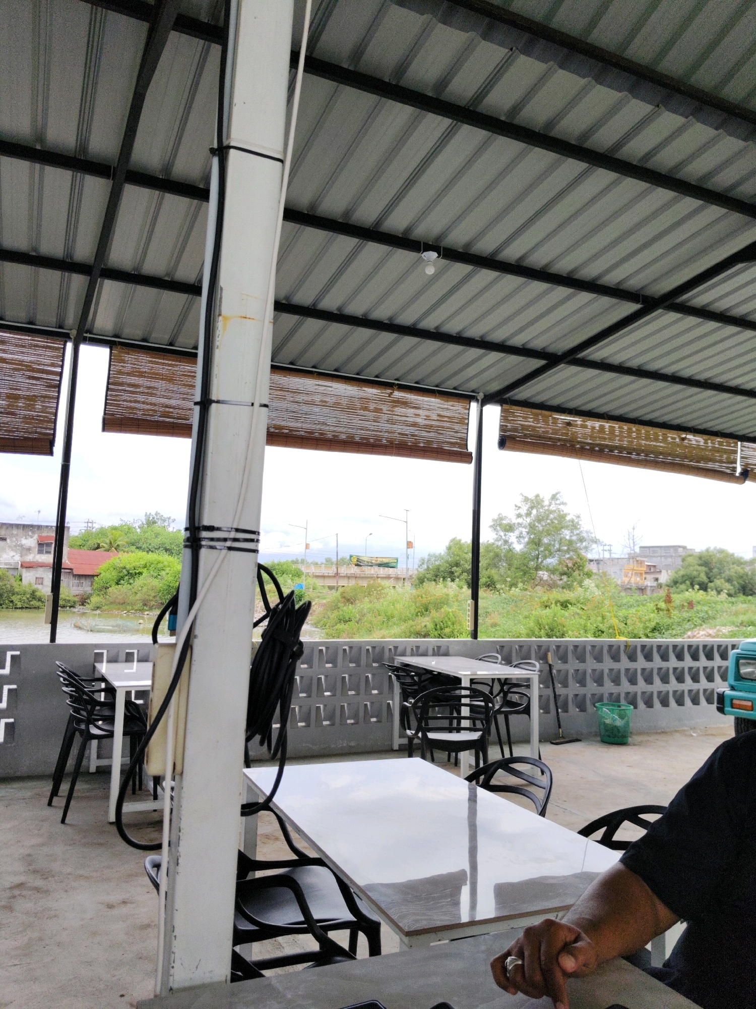 The image depicts an outdoor seating area of a restaurant or cafe, likely located in Lhokseumawe, Indonesia, given the context. The area is covered by a corrugated metal roof supported by metal beams. Bamboo blinds are visible, serving as partial walls. In the foreground, a person's arm and hand wearing a ring are resting on a table. Several tables and chairs are arranged in the seating area. In the background, there is a body of water, lush greenery, buildings, and infrastructure, suggesting an urban or semi-urban environment. A turquoise vehicle is partially visible on the right side of the image. The overall impression is a casual outdoor dining or resting space.