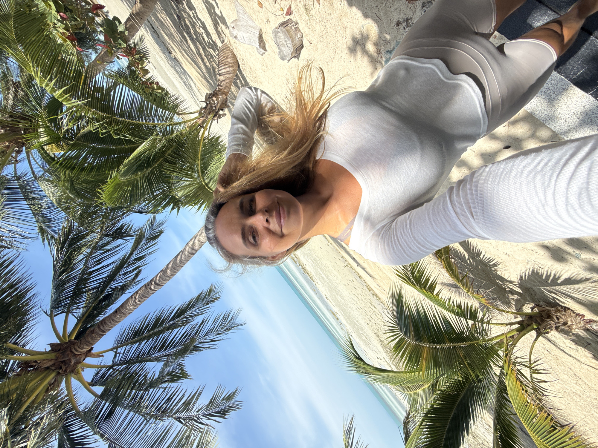 In Ko Samui District, Thailand, a fair-skinned woman with blonde hair is taking a selfie on a sandy beach. She is wearing a white long-sleeved shirt and beige biker shorts. Palm trees with green fronds are visible in the foreground and background, with the ocean and a clear blue sky also present. The woman is holding her hair with her left hand, and her right arm is extended, holding the camera. The lighting suggests it is daytime, and shadows from the palm trees are cast on the sand. Some scattered rocks or driftwood are visible on the beach.