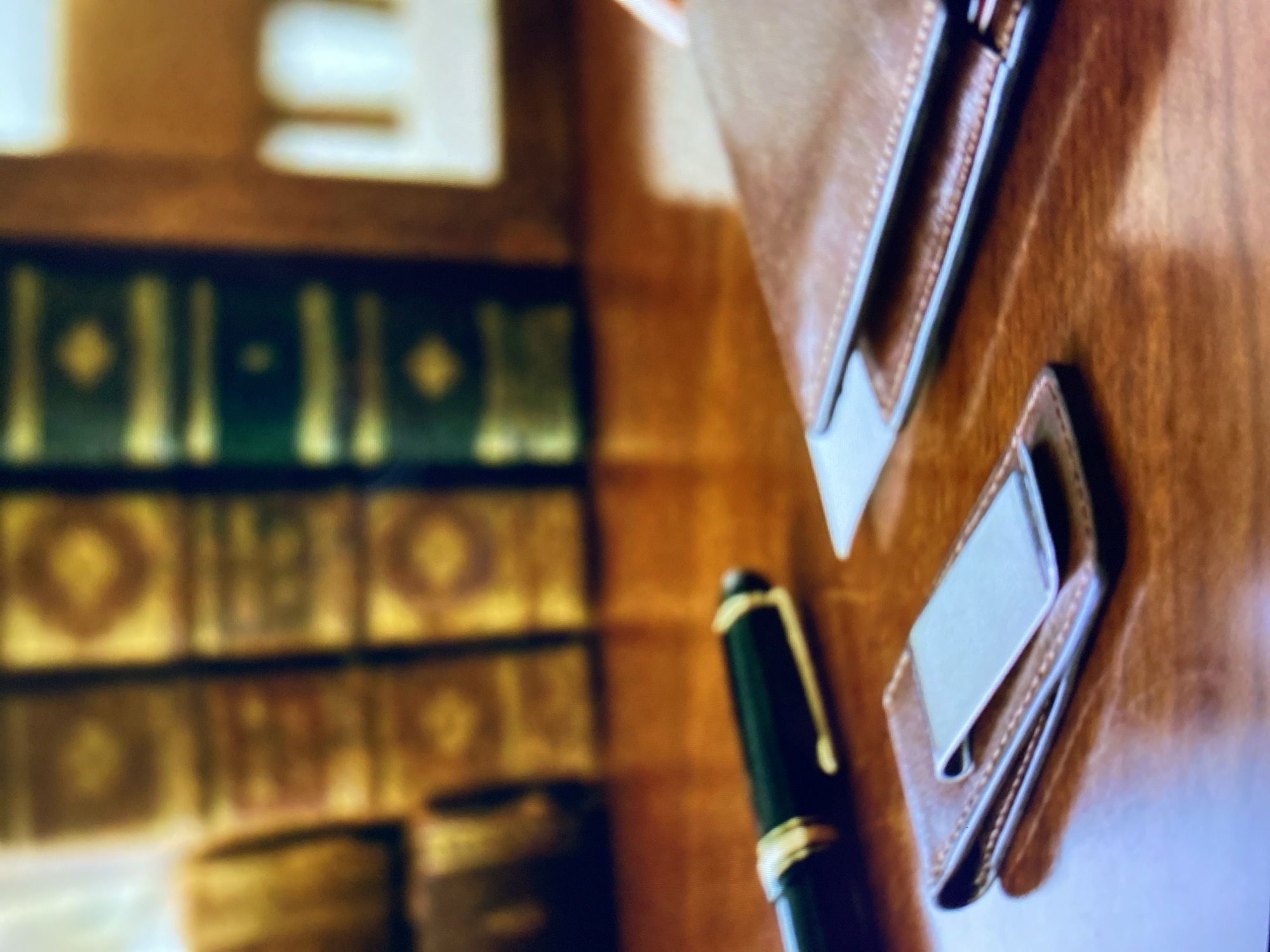 The image shows a close-up, eye-level, medium shot of a wooden desk surface with various stationery items and books in the background. In the foreground, a black fountain pen with gold accents lies horizontally on the desk, pointing towards the left. To the right of the pen, a brown leather cardholder or wallet is placed, with a portion of a white card peeking out from it. A silver money clip is positioned on top of the cardholder. In the background, several books with ornate gold detailing on their spines are visible, arranged on what appears to be a bookshelf. The overall lighting suggests an indoor setting, possibly an office or study. The image does not contain any information about the city of Kuta Selatan, Indonesia.