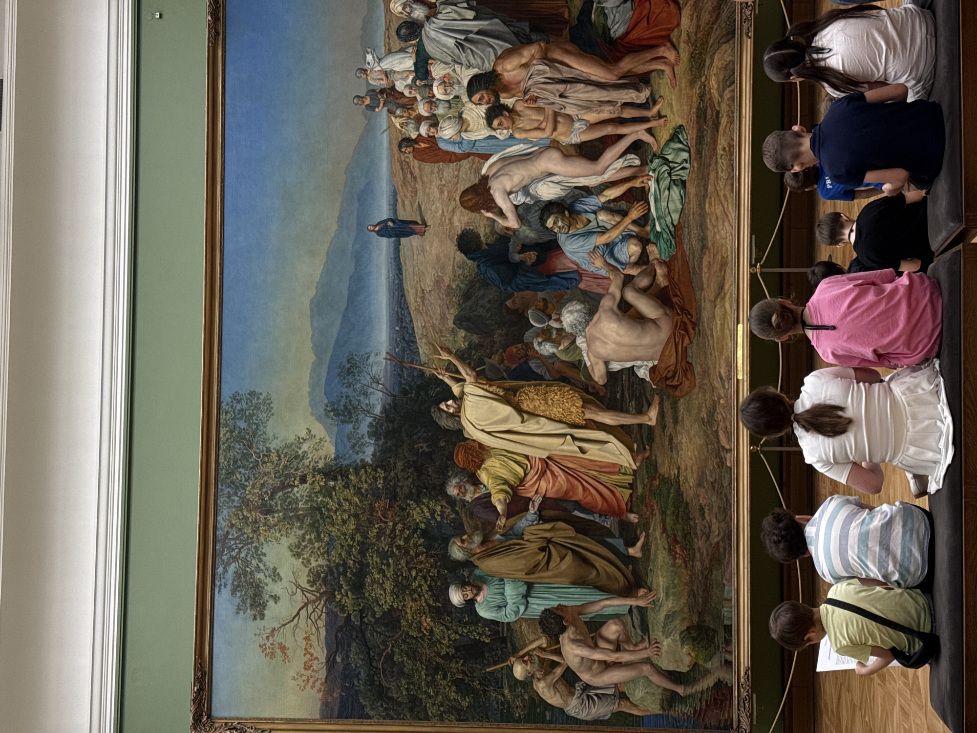 The image shows a group of children seated on a bench, viewing a large oil painting inside what appears to be a museum or gallery. The painting is prominently displayed with a gilt frame. It depicts a large group of figures, including nudes, in a scene with trees and mountains. The painting's style suggests a classical or historical subject. The setting might be in Moscow, Russia, although this is not explicitly confirmed in the image.