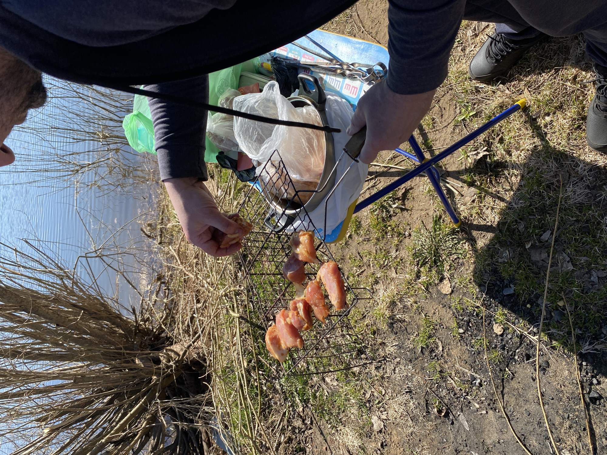 A partially obscured individual prepares raw meat for grilling outdoors next to a body of water, possibly near the city of Noviy, Russia. The person, wearing gray long sleeves and dark pants with black sneakers, places the meat on a metal grill. A blue folding table with a light blue top supports bags of food and cooking utensils, including skewers and a pan. The environment is grassy with bare patches of soil and some remnants of burnt material, indicative of past fires. Branches and a body of water are visible in the background.
