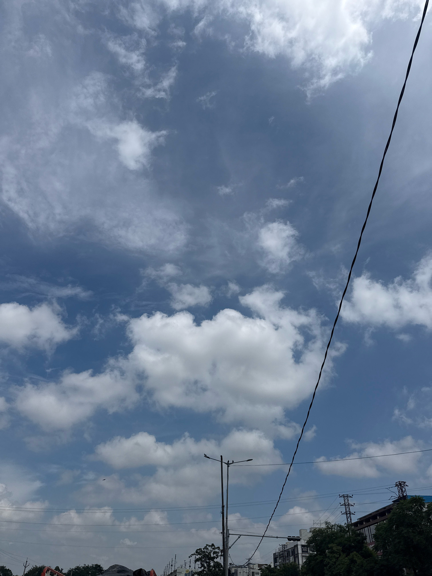 The image depicts a cloudy sky over a city with a prominent power line running diagonally from the top right to the bottom left. In the lower portion of the image, a street lamp and several buildings are visible. The sky is a mix of blue and white clouds, with some areas appearing darker, suggesting overcast conditions. The power line is black and appears to be a single wire. The buildings are of various heights and architectural styles, with some featuring windows and balconies. The street lamp is a typical urban design with a single light fixture. The overall scene suggests an outdoor environment in a populated area, likely during the daytime due to the brightness of the sky. The presence of the power line and buildings indicates an urban setting, and the specific reference to Hyderabad, India, contextualizes the location.