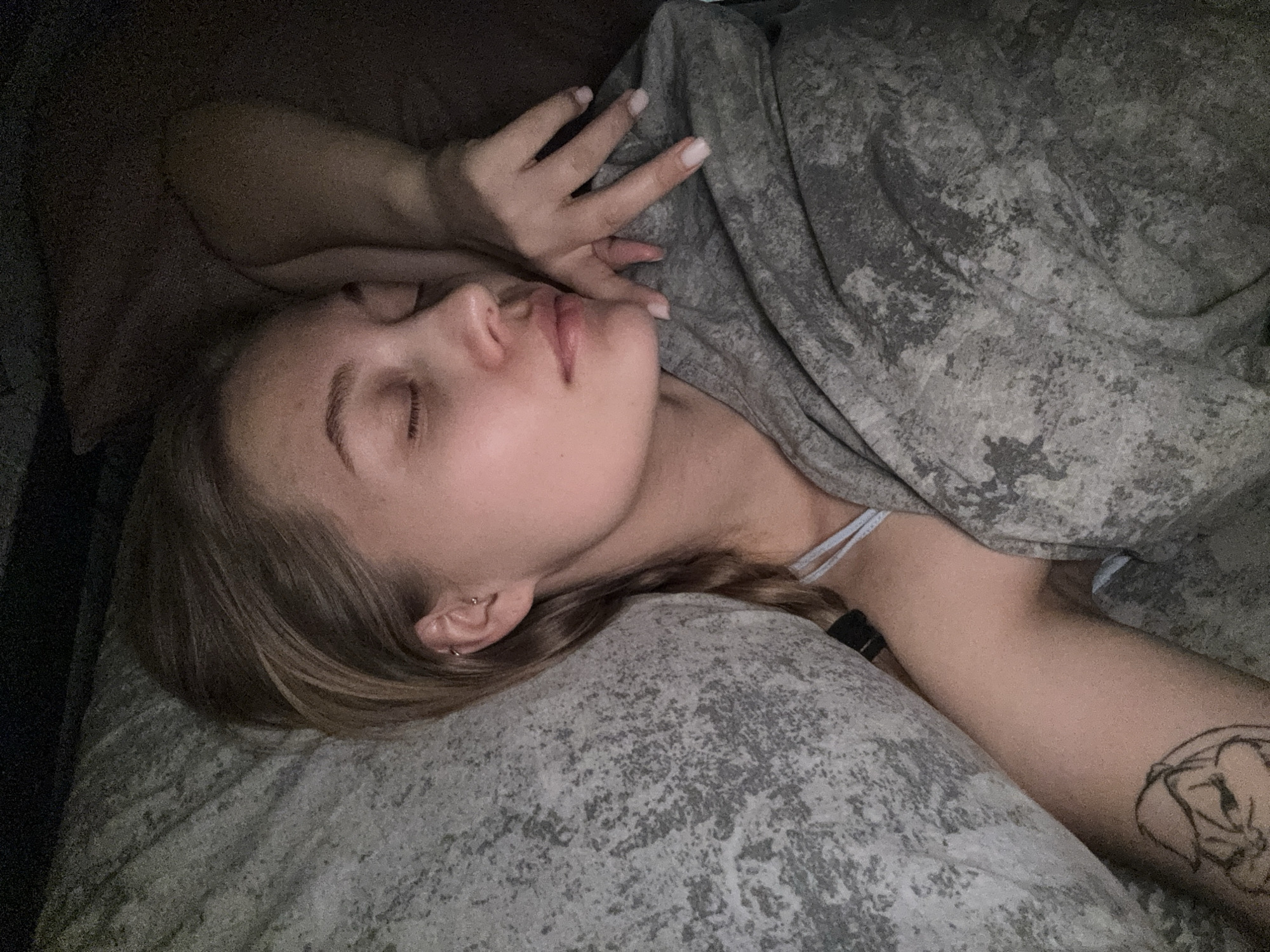 A young person, likely female, is depicted sleeping or resting peacefully indoors, presumably in a bed in Chaikovsky, Russia. She is lying on her side, facing slightly away from the viewer, with her eyes closed and a calm expression. Her right hand is gently placed near her head, resting on the pillow, while her left arm is extended under a blanket.
She has light brown hair, which appears long and somewhat gathered or braided. Visible details include a small hoop earring in her earlobe and another small piercing higher up on her ear. A light-colored strap, possibly from a bra or a camisole, is visible near her shoulder. On her right arm, a black outline tattoo is visible, depicting a stylized figure or object. A dark band, possibly a watch or fitness tracker, is on her left wrist.
The setting appears to be a bedroom, characterized by dim lighting, suggesting it is nighttime or a darkened room. The blanket and pillow both feature a mottled, abstract grey and black pattern. The overall atmosphere conveys tranquility and rest. No text is visible in the image.