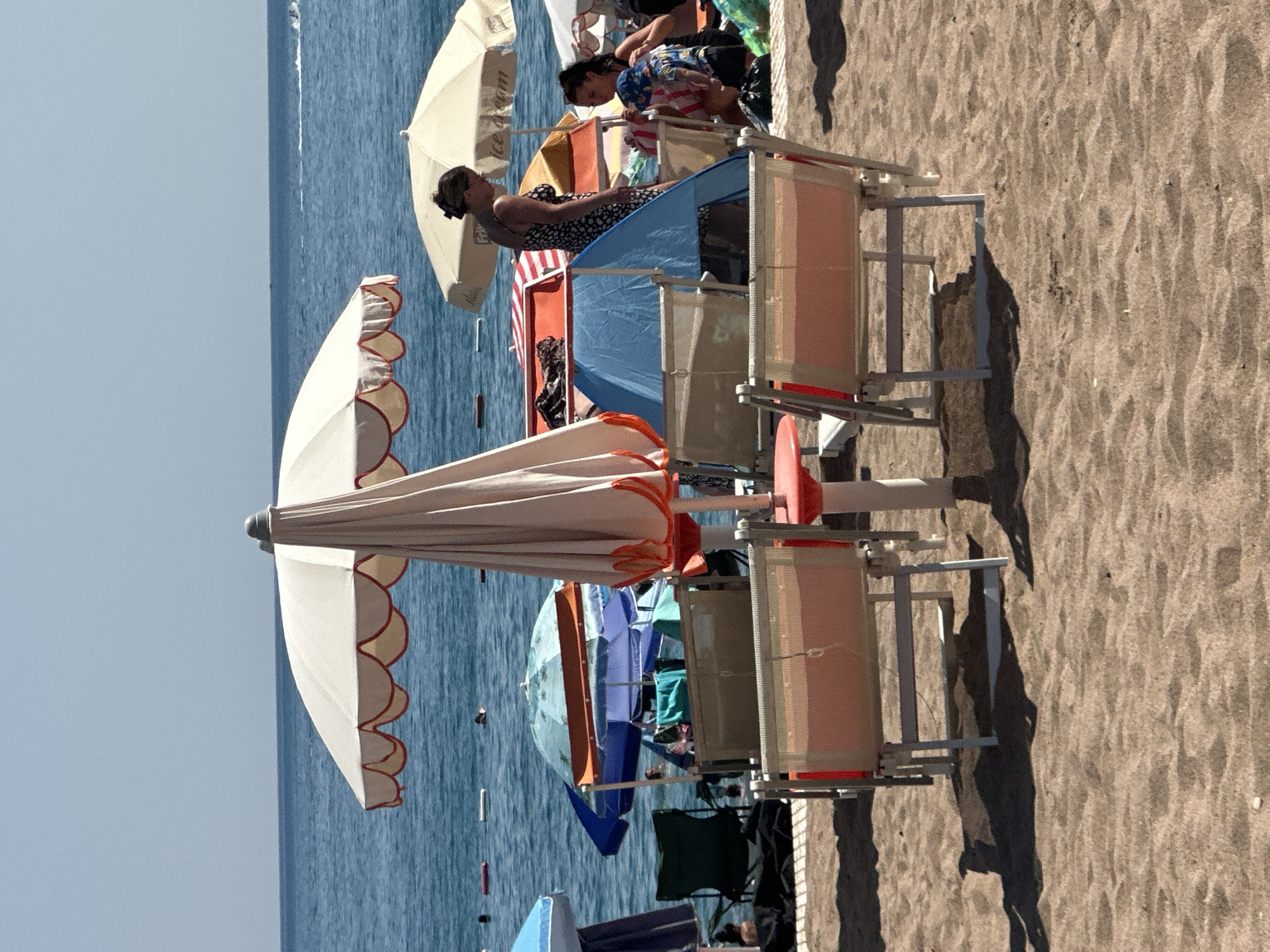The image depicts a sandy beach with a calm blue sea in the background. Several beach umbrellas are set up, with some people visible enjoying the sunny weather. In the foreground, two empty beach chairs are positioned on the sand. A woman in a patterned dress is standing near a cream-colored umbrella, and a smaller child in a blue hat is with another person in the background. The overall scene suggests a relaxed day at the beach in Boreti, Montenegro.