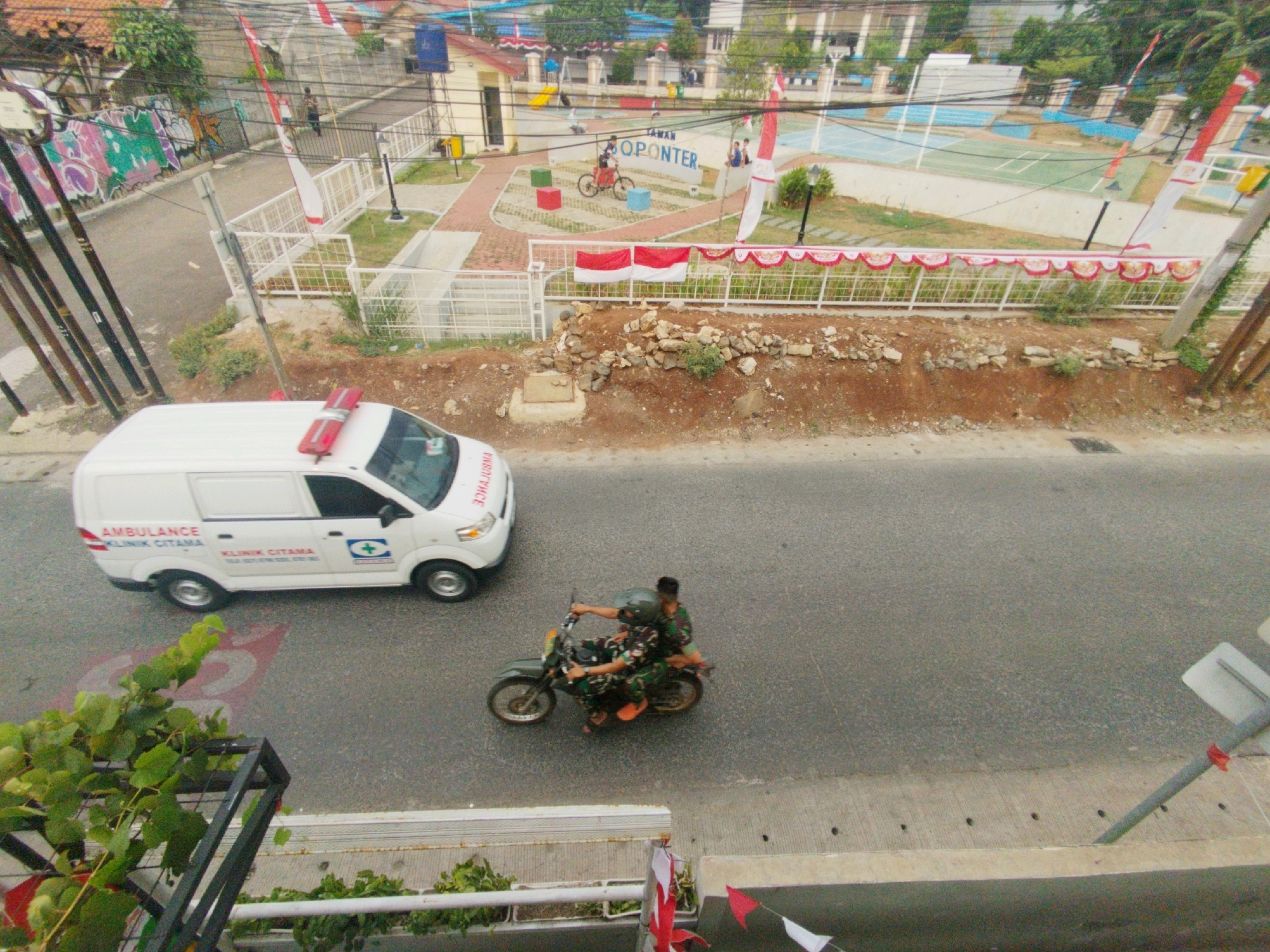 The image shows a street scene in Depok, Indonesia, with an ambulance and two people on a motorcycle. The ambulance is white with red lights on the roof and "AMBULANCE KLINIK CITAMA" written on its side. The motorcycle is dark green and carries two people in military-style camouflage uniforms. One person is driving the motorcycle, and the other is riding behind them. The street is asphalt, and there are some red markings on it. In the background, there are buildings, trees, and a park with a basketball court. The park has a sign that says "TAMAN OPONTER." Indonesian flags are visible on poles and decorations. The overall impression is a busy street scene in a residential or commercial area.