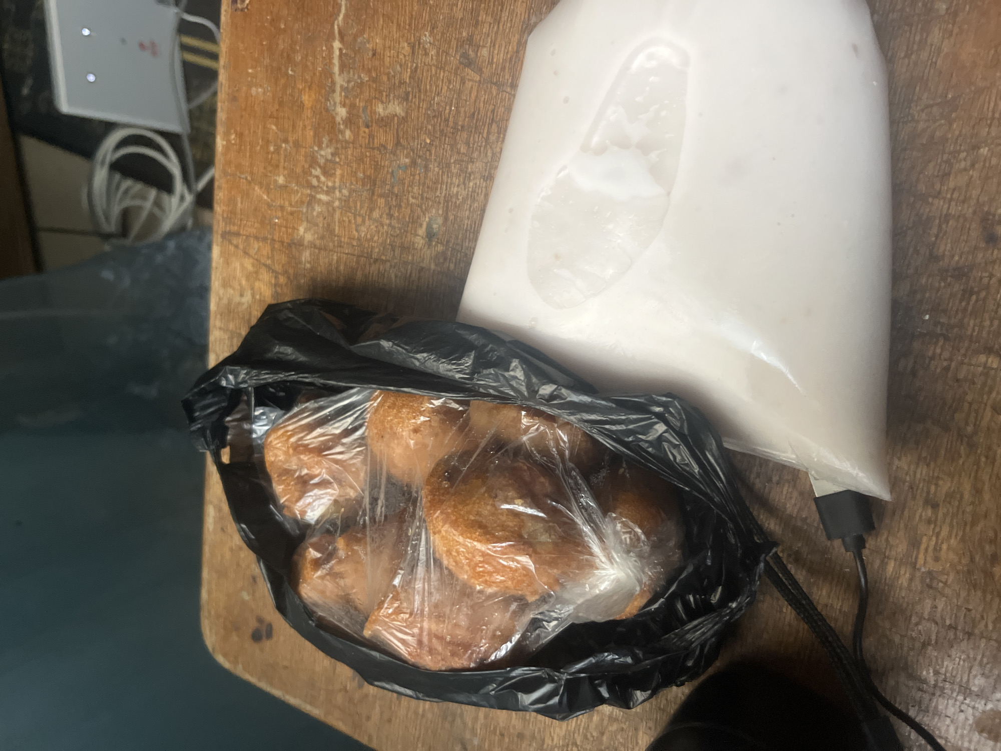 The image shows a loaf of bread in a black plastic bag, and a bag of liquid, possibly soup or sauce. They are on a brown wooden surface. The scene is consistent with a domestic setting in Barakin Akawo, Nigeria, based on the food packaging and the general environment. There are no people visible in the image.