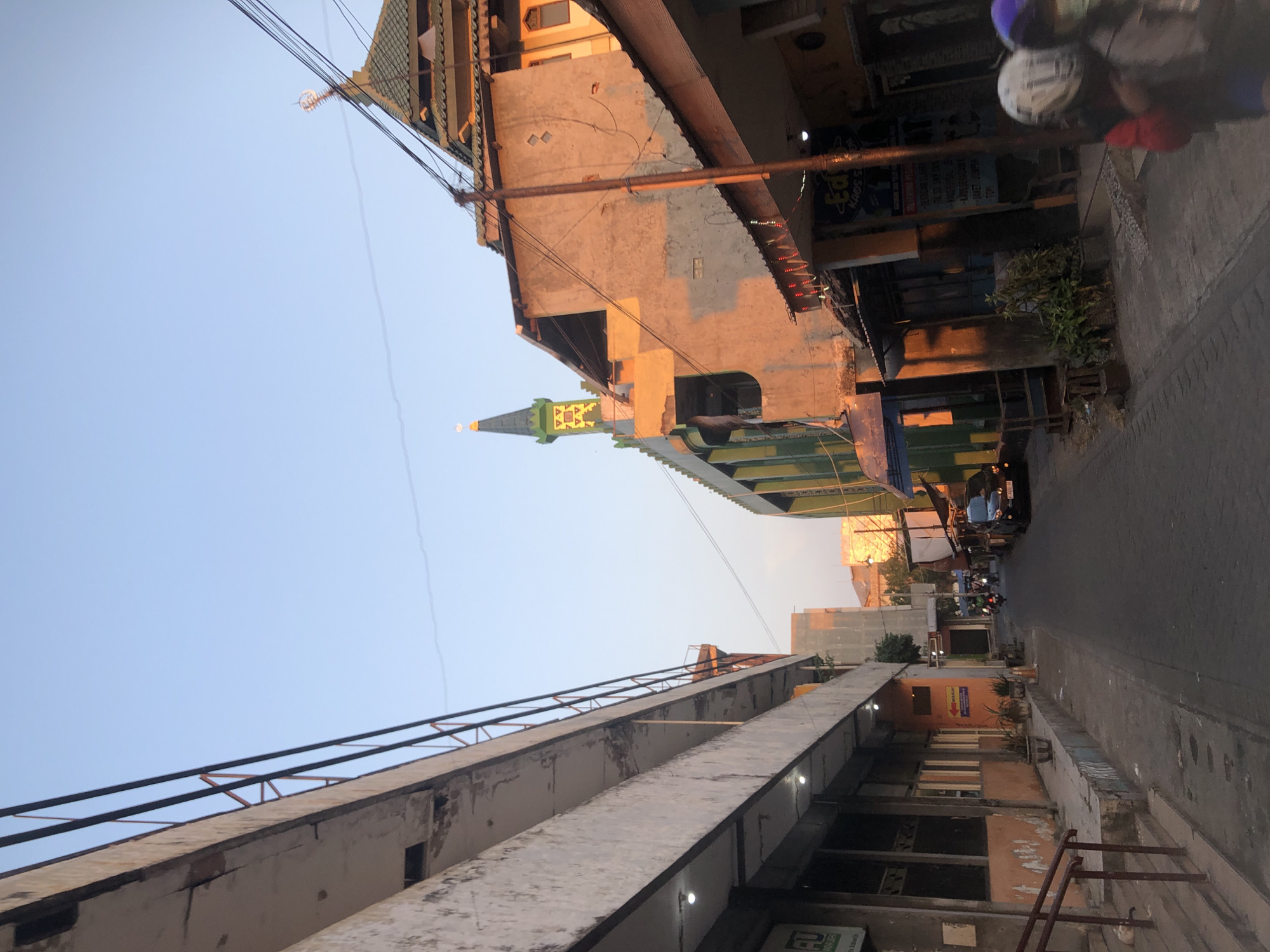 The image depicts a narrow street in Sidoarjo, Indonesia, bathed in the warm glow of the setting sun. On the left, a weathered, multi-story building with a concrete facade features an elevated walkway supported by metal beams. The building's lower level appears to house businesses, indicated by shopfronts and illuminated by artificial lights. To the right, a mosque with a green, tiered roof and a prominent minaret stands out against the clear blue sky. Adjacent to the mosque are several smaller buildings with shops, their facades illuminated by the sun, casting long shadows. A black car is parked on the street, and several motorcycles are visible in motion, with riders wearing helmets. The street itself is paved, and potted plants are placed along the sidewalk. The scene captures a typical urban environment in Indonesia during the late afternoon or early evening.