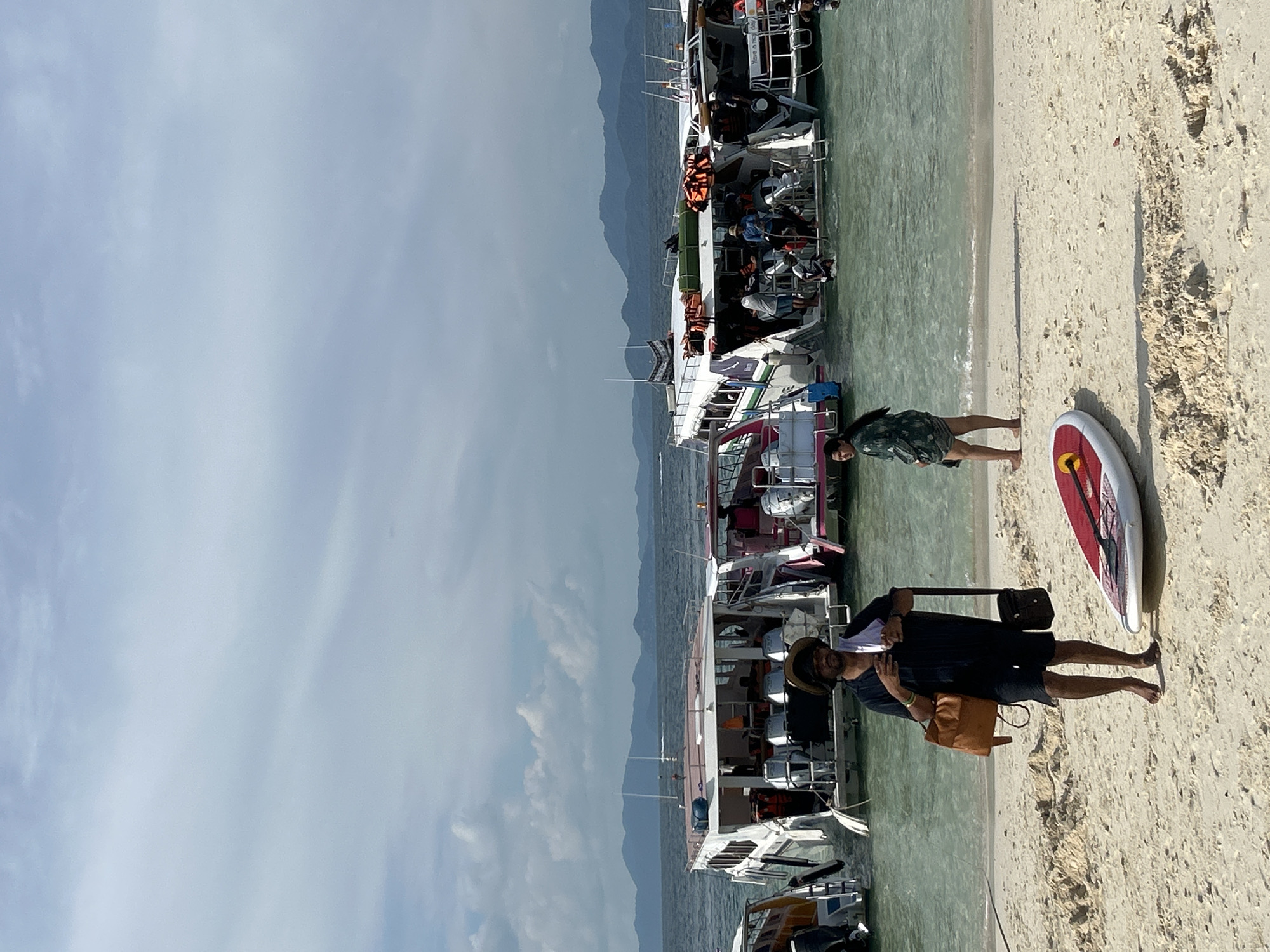 In Tambon Phru Nai, Thailand, a sandy beach with a clear, shallow ocean is the setting for a scene with several boats and people. In the foreground, a man wearing a hat, dark shirt, and shorts stands on the sand. He has a brown bag over his right shoulder and a smaller dark bag hanging from his left. Next to him, a red and white paddleboard lies on the sand. A woman in a patterned shirt and shorts stands facing away from the camera, looking towards the water. In the middle ground, several passenger boats are anchored close to the shore. The boats are white with colorful accents and are carrying groups of people. Some individuals are standing on the boats, while others are in the shallow water, possibly disembarking or preparing to board. The water is a translucent turquoise color, revealing the sandy bottom in places. In the background, rolling hills covered in lush greenery rise from the sea, under a sky with scattered clouds. The overall atmosphere suggests a popular tourist destination for island hopping or water activities.