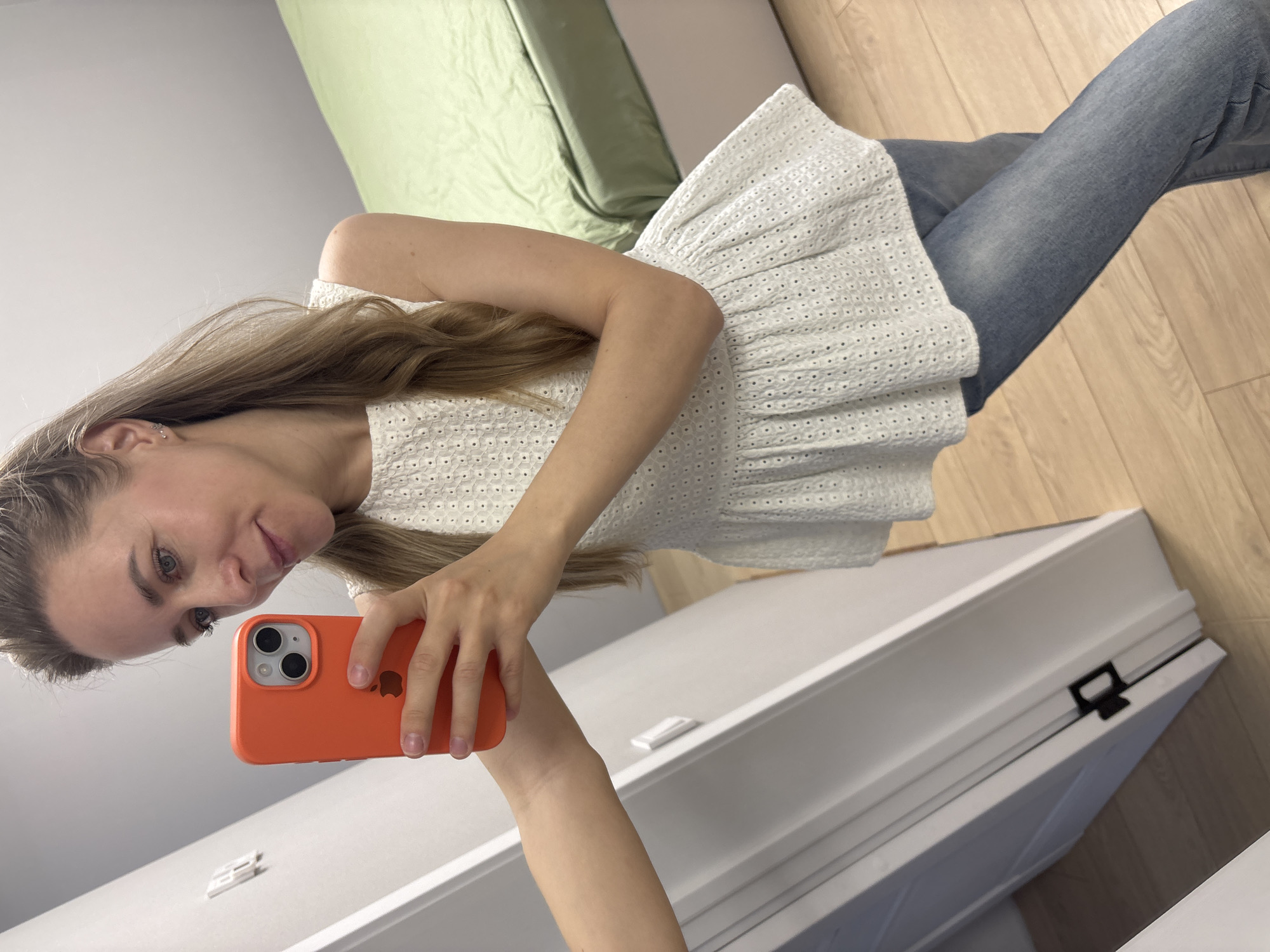 A fair-skinned woman with light blonde hair is taking a selfie with an orange iPhone. She is wearing a white, sleeveless eyelet top and blue jeans. The background includes a light green cushion or piece of furniture, a white door or cabinet, and a light wood floor. The location is possibly Chaikovsky, Russia, but this is based on contextual information external to the image.