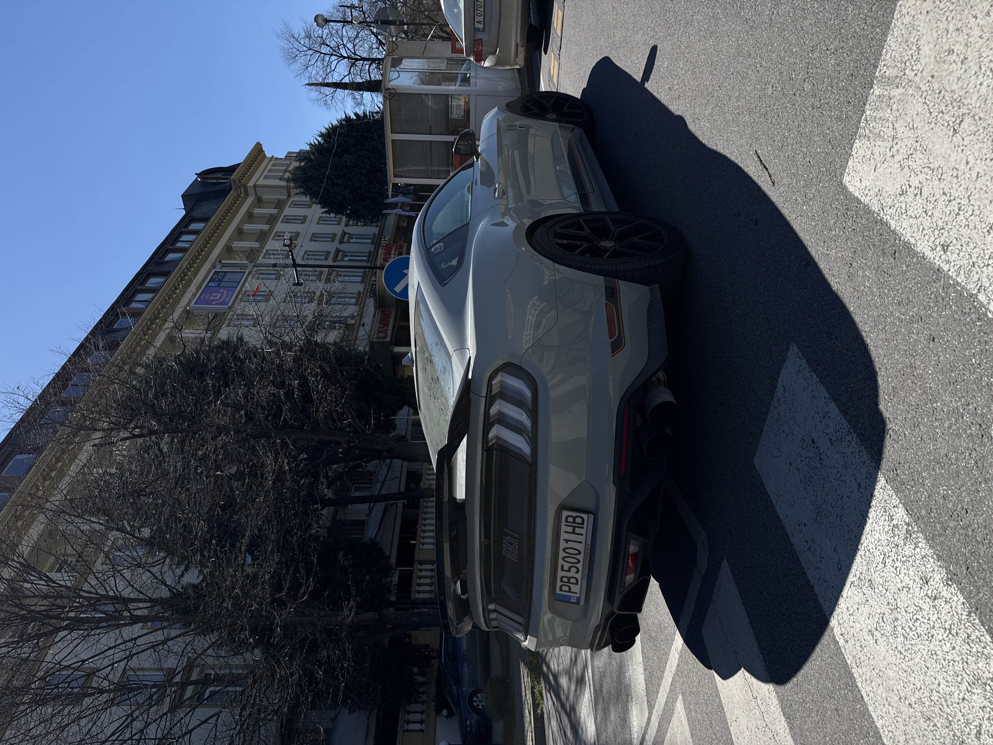 A gray Ford Mustang Mach 1 is parked on a pedestrian crosswalk in Plovdiv, Bulgaria. The car's license plate reads "PB5001HB". The background shows a multi-story building with shops, and bare trees line the street. The sky is clear and blue. Other vehicles are present in the scene.