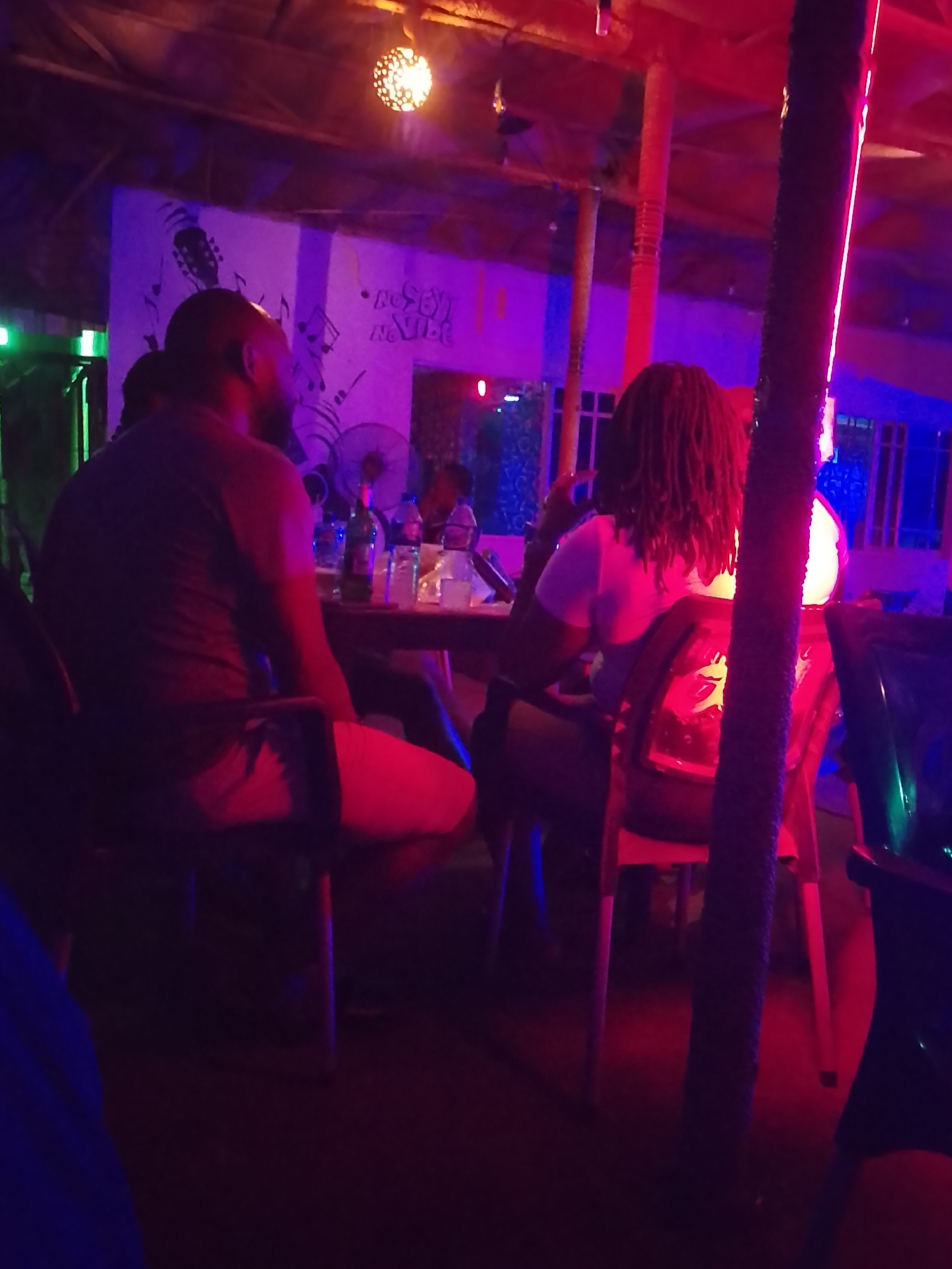 Inside a bar in Unknown, Unknown, two people are sitting at a table. One person is wearing a grey shirt and red shorts, while the other is wearing a white shirt. The bar has a mural on the wall, and there are several bottles and glasses on the table. The lighting is dark, with red and blue lights creating a nightclub vibe.