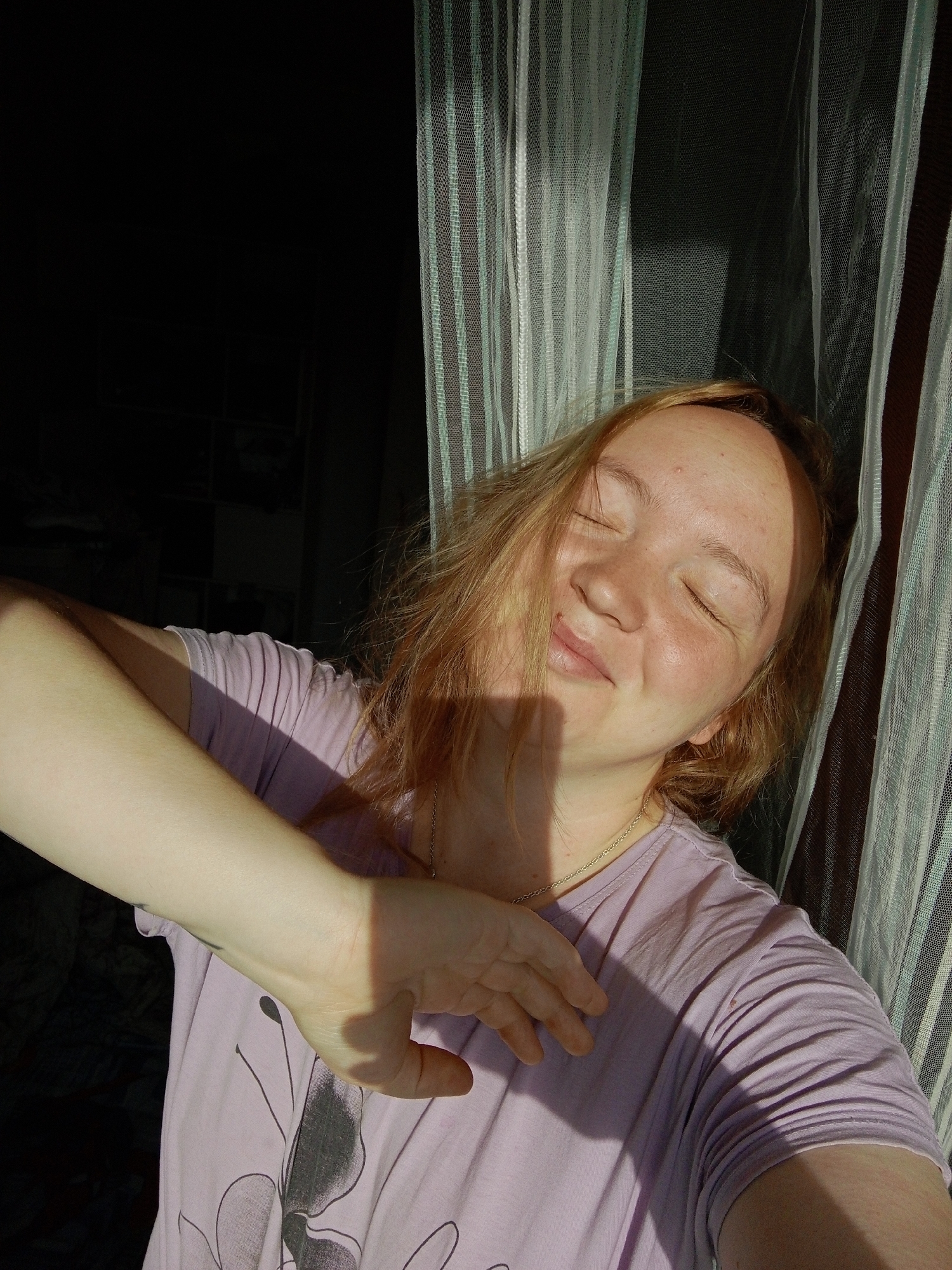 A young adult female is posing for a selfie. She has light skin, reddish-blonde hair, and her eyes are closed. She is wearing a light purple t-shirt and a gold necklace. Her right arm is extended, casting a shadow on her face and shirt. The background includes sheer, striped curtains, and a dark, undefined space. The location is unknown.