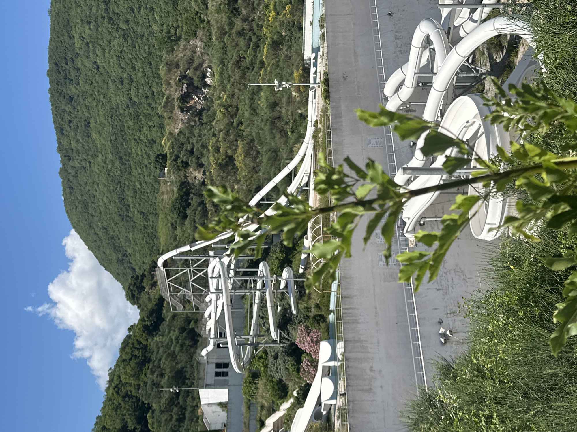 The image depicts an abandoned water park nestled on a hillside in Podostrog, Montenegro. The main features are several white plastic water slides, some of which are suspended on metal scaffolding, winding their way down the slope. The slides appear to be empty and unused, with some sections showing signs of wear and tear. In the background, lush green trees and vegetation cover the hillside, with a clear blue sky overhead. A large, white building structure is visible near the top of the slides, and other smaller structures and concrete barriers are scattered throughout the scene. The overall impression is one of a once-vibrant recreational facility now in a state of disrepair, reclaimed by nature. There are no people visible in the image.