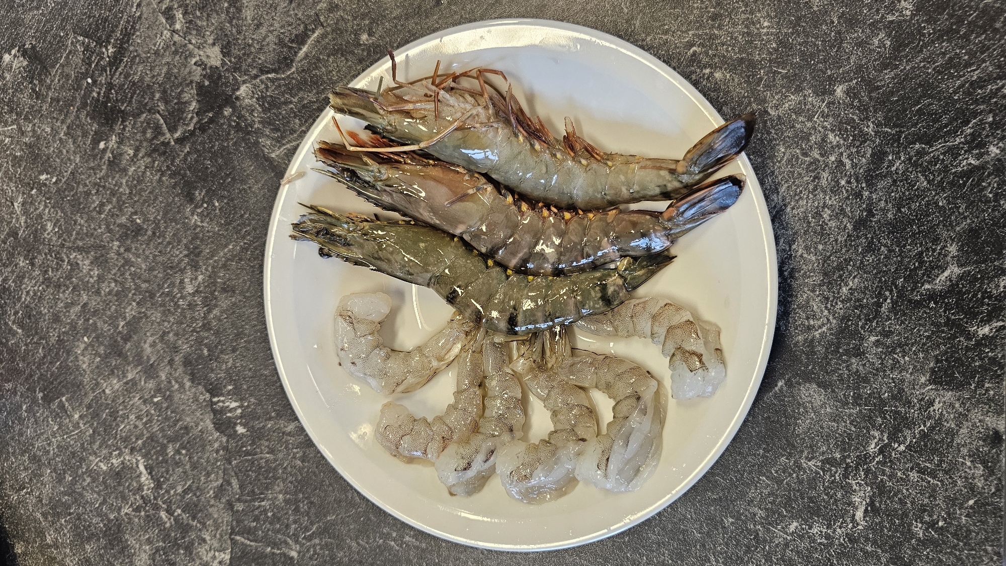A white, round plate, centered on a dark, mottled surface, holds a selection of raw shrimp. On the upper half of the plate, three whole, unpeeled shrimp are visible, characterized by their grey-green bodies, dark banding, and intact heads with long antennae. Below these, approximately six to seven peeled and deveined shrimp tails are arranged, appearing translucent white with subtle pinkish tones. The shrimp are uncooked and appear fresh, likely prepared for a meal. The background surface is a dark, grayish-black with irregular lighter patterns, resembling a stone countertop. There are no people, visible activities, specific time of day indicators, or discernible text in the image. While the location context is Philippeville, Belgium, no visual cues within the image confirm this.