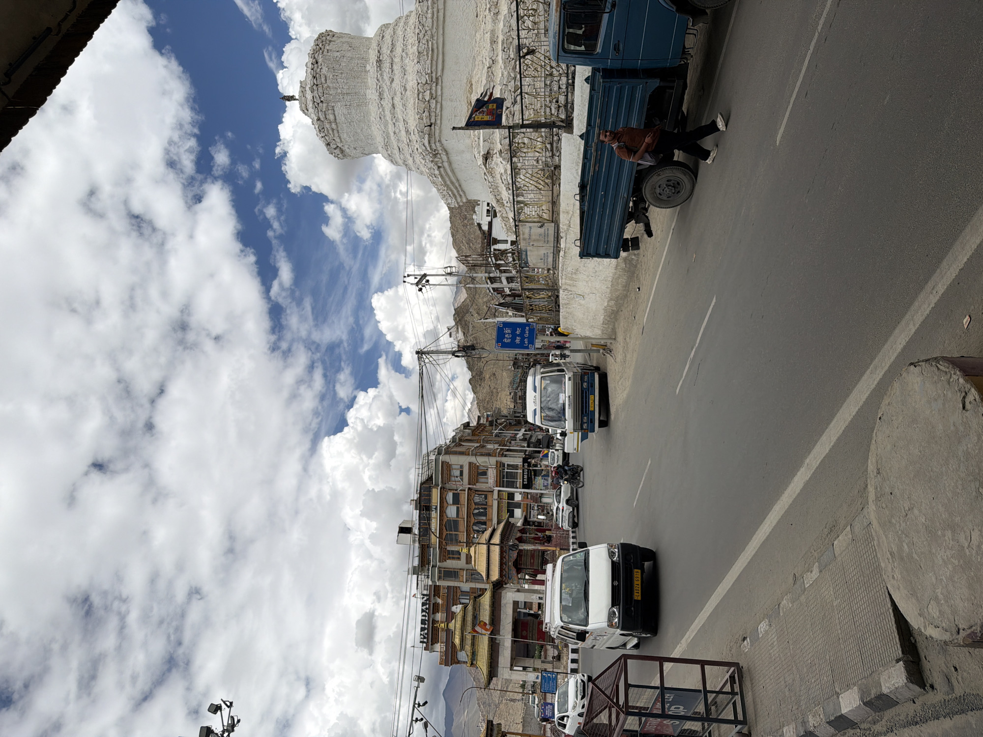 In the city of Leh, India, a street scene unfolds under a cloudy sky with patches of blue. Dominating the right side of the frame is a large, white stupa, a Buddhist religious monument, with a figure standing atop its peak. Adjacent to the stupa is a blue truck, and a person in dark pants and a reddish-brown shirt walks past it on the road. The road is lined with traffic, including a white van with a yellow license plate, a white car, and a bus. Buildings line the street, featuring traditional Ladakhi architecture with golden roofs and intricate designs. Traffic signals and utility poles are visible, indicating an urban setting. A pedestrian walkway with a metal barricade is present on the left side of the road. The overall impression is a bustling street in Leh, a popular tourist destination known for its monasteries and stunning landscapes.