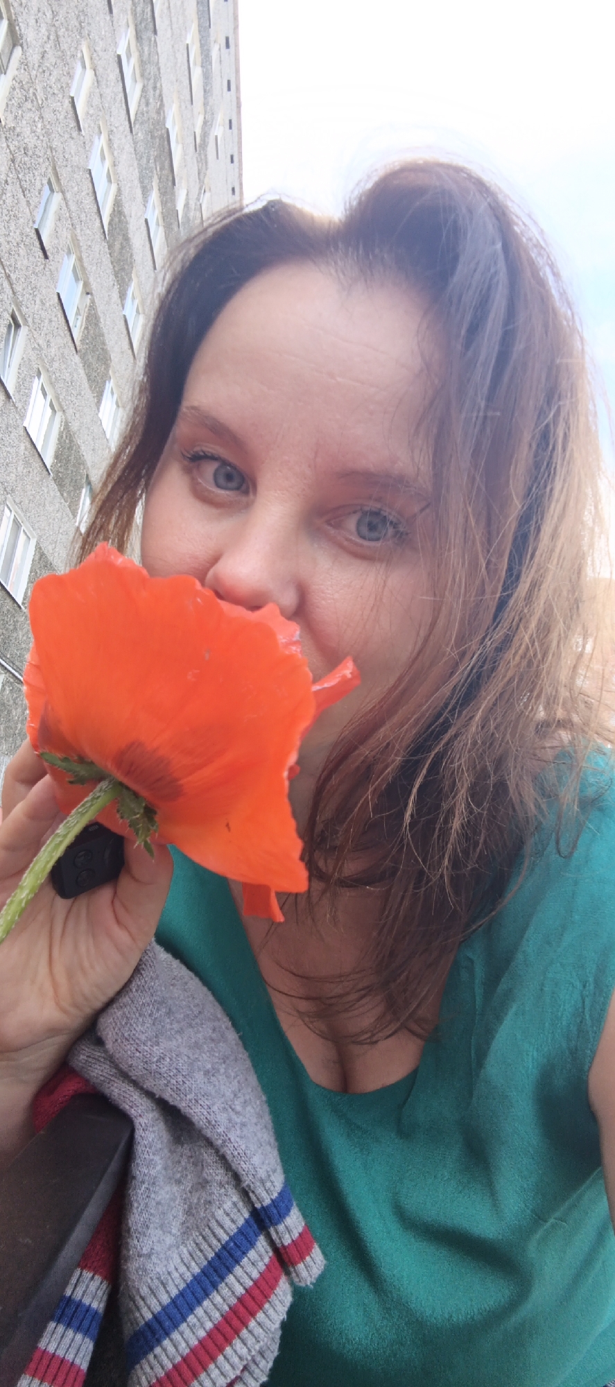 A woman with light skin and brown hair holds an orange poppy in front of her face. She is wearing a teal top and a gray cardigan with red and blue stripes. A building with many windows is visible in the background. The woman is looking at the camera and smiling. The scene appears to be outdoors, possibly in a residential area of Chaikovsky, Russia, given the architecture of the building.