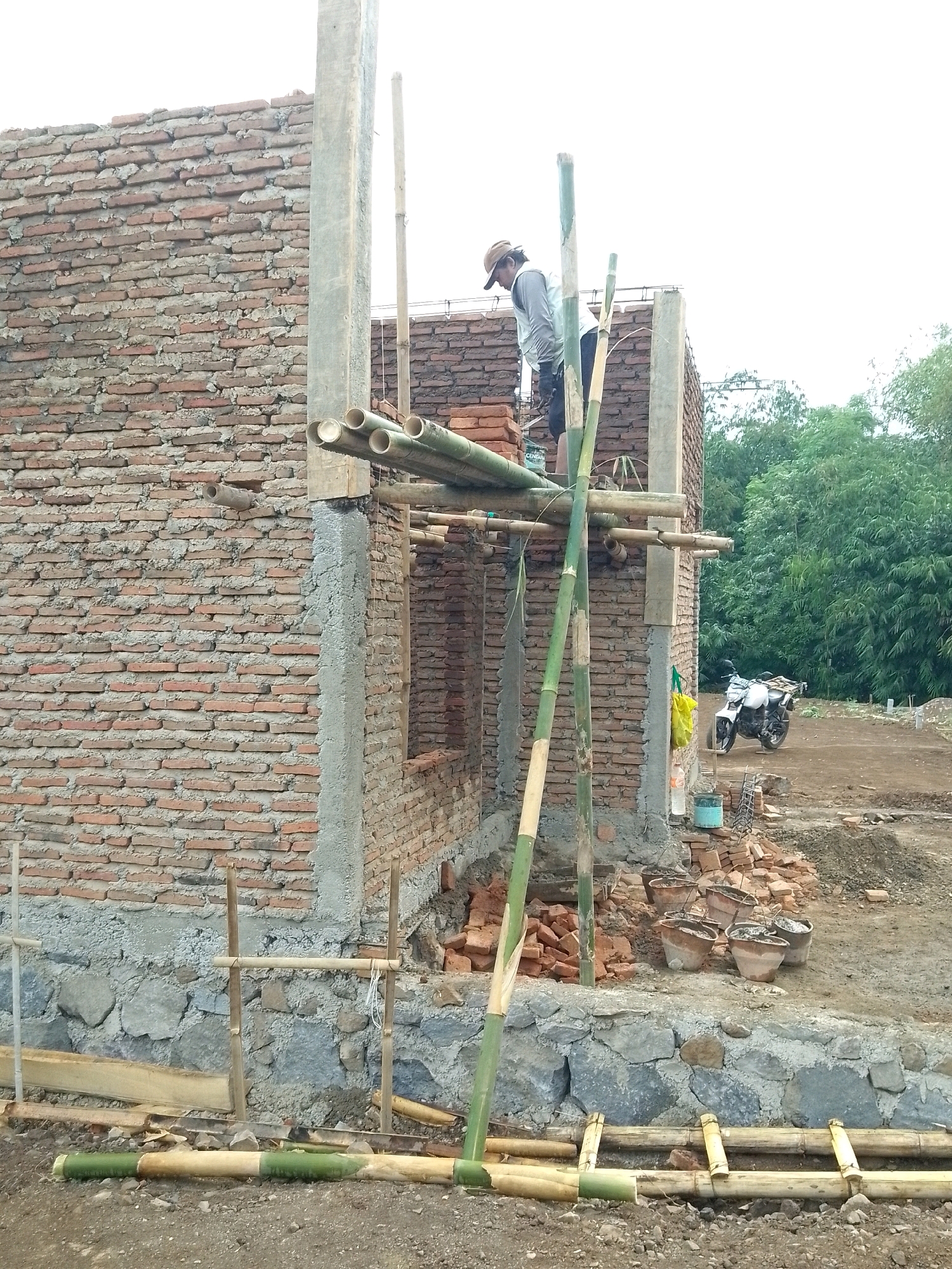 A construction scene unfolds in Jember, Indonesia, where a brick building is under construction. A construction worker, wearing a hat and a light-colored vest over a dark shirt, stands on a temporary scaffold made of bamboo. The scaffold supports bundles of bamboo pipes. The building features brick walls with visible mortar, and a concrete pillar is present. In the background, a white motorcycle is parked on a dirt path amidst lush green trees. Several buckets and piles of bricks are scattered around the construction site. The ground is composed of dirt and gravel.