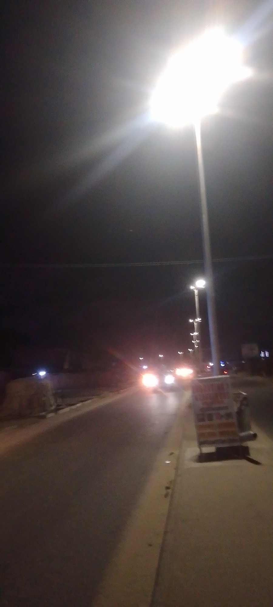 The image depicts a nighttime street scene in the city of Unknown, Unknown. A dark asphalt road extends into the distance, bordered on the right by a sidewalk or verge. A tall streetlamp on the right foreground emits a brilliant white light, causing significant lens flare across the upper right quadrant. Further along the right verge, a rectangular object, possibly a mobile vendor stand or sign, displays reddish and white elements. Multiple vehicles approach in the mid-ground, identifiable by their bright white and yellow headlights. Faint red lights, potentially taillights, are visible further down the road. On the left, dark shapes represent distant structures or natural elements, with a lighter, possibly concrete, barrier visible closer to the road. Additional streetlights are present along the road's length. Faint horizontal lines, potentially power lines, traverse the dark sky. No individuals are visible.